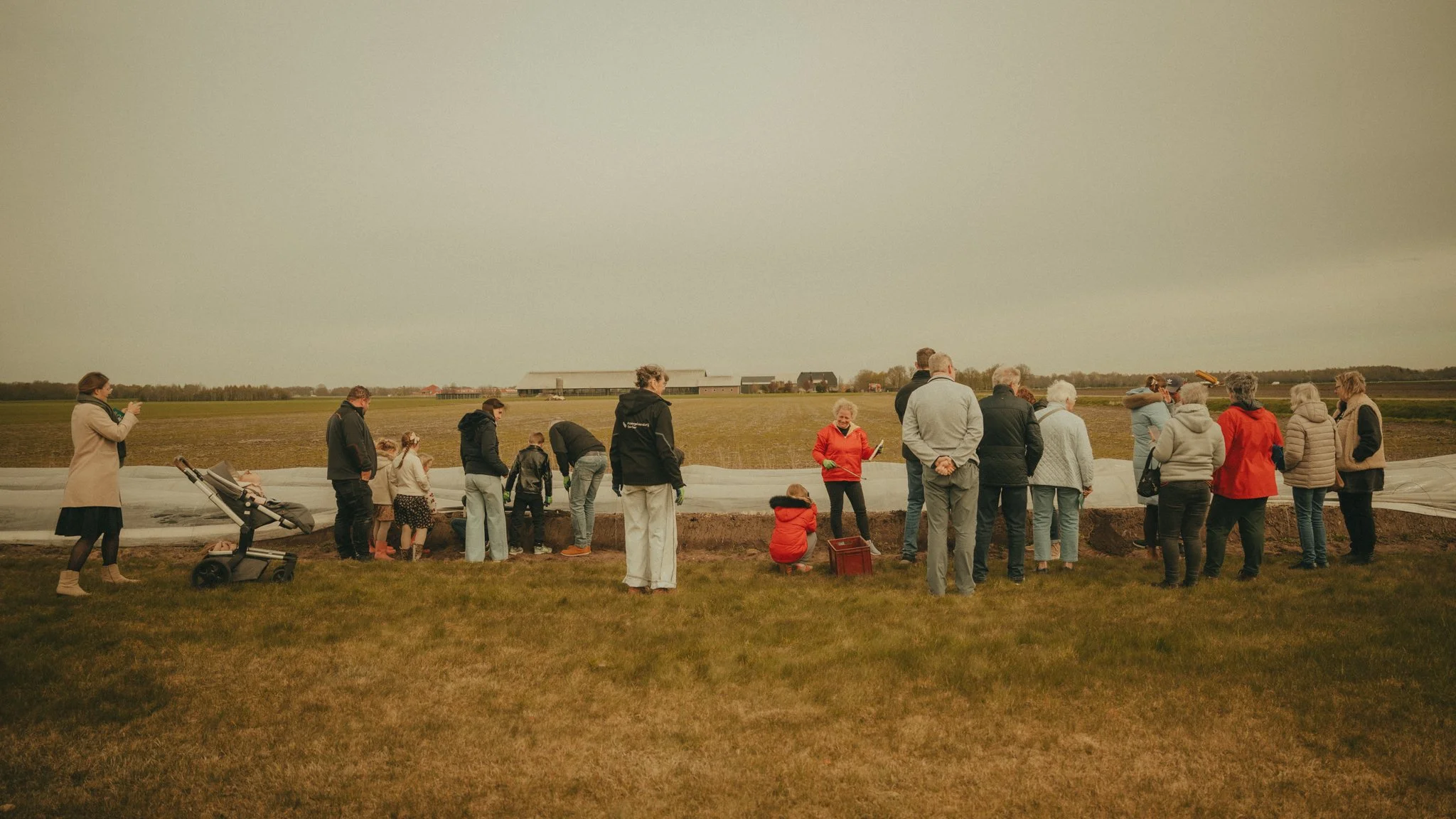 A group of people gathered outdoors, observing an event, with a cloudy sky and open field in the background.