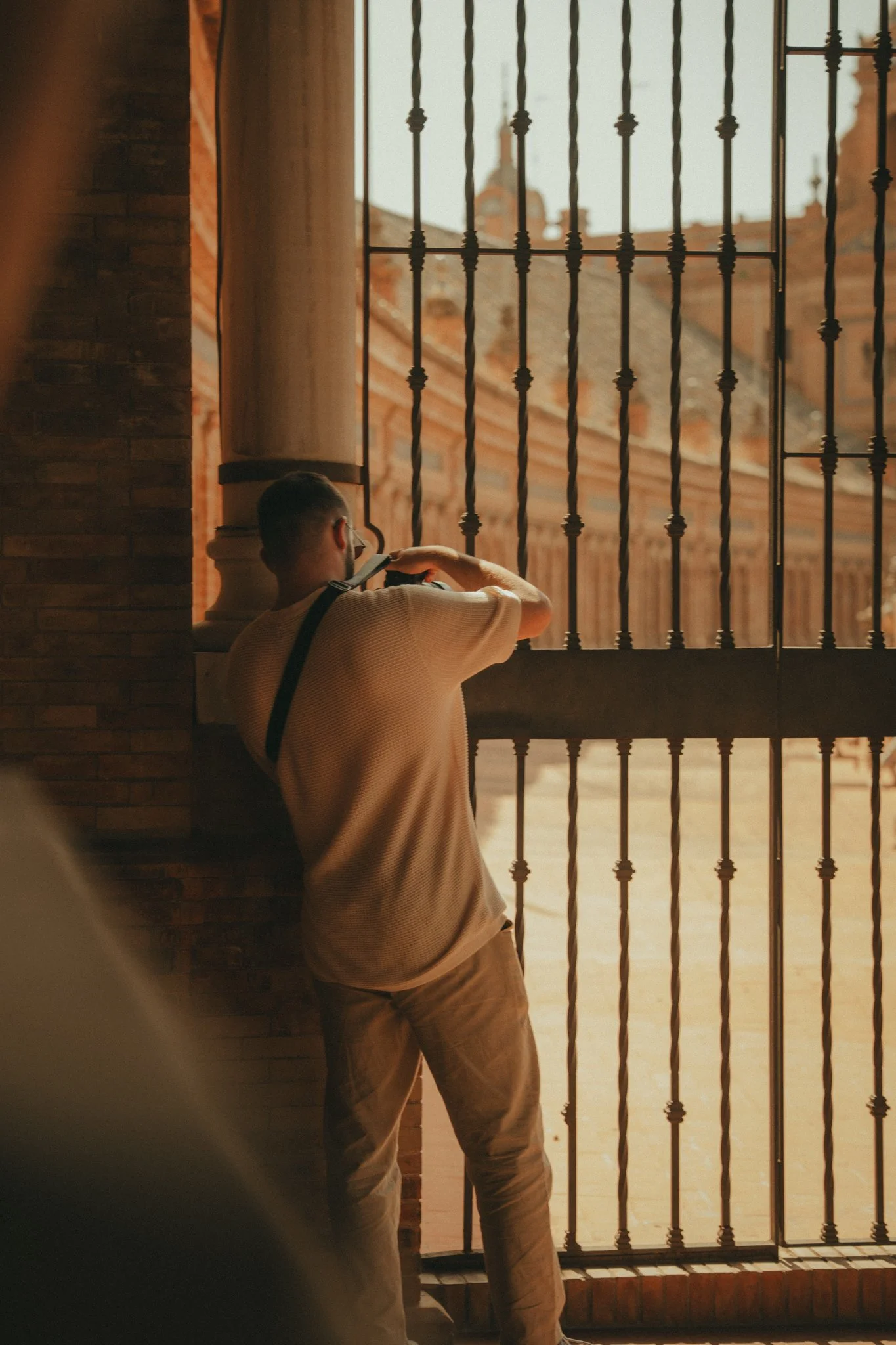 A man leaning against a brick wall near a metal gate, looking out at buildings with rooftops. The scene is lit with warm sunlight.