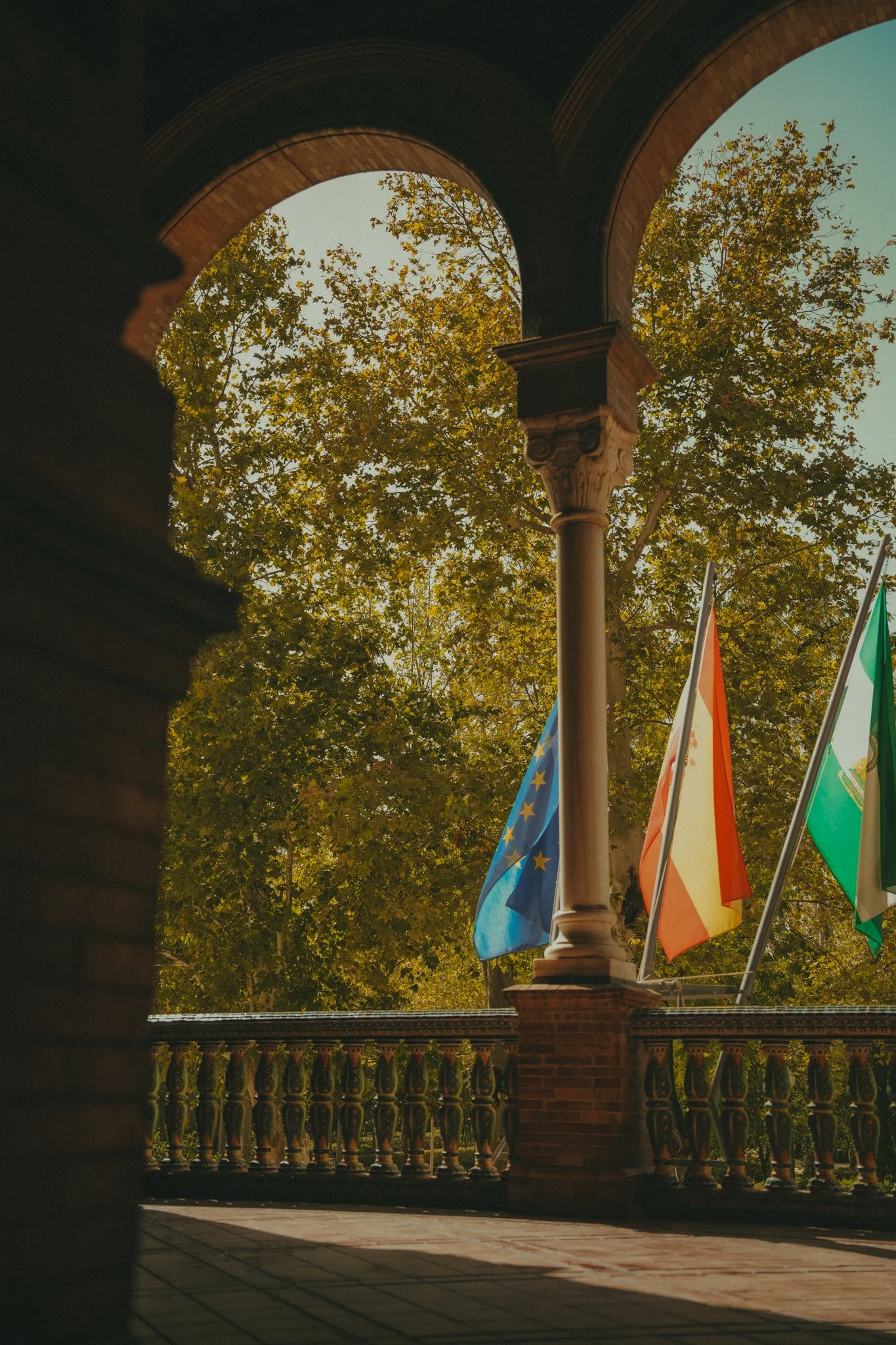 View of flags from different countries, seen through an archway with a decorative balustrade and brick columns, with trees in the background during daytime.