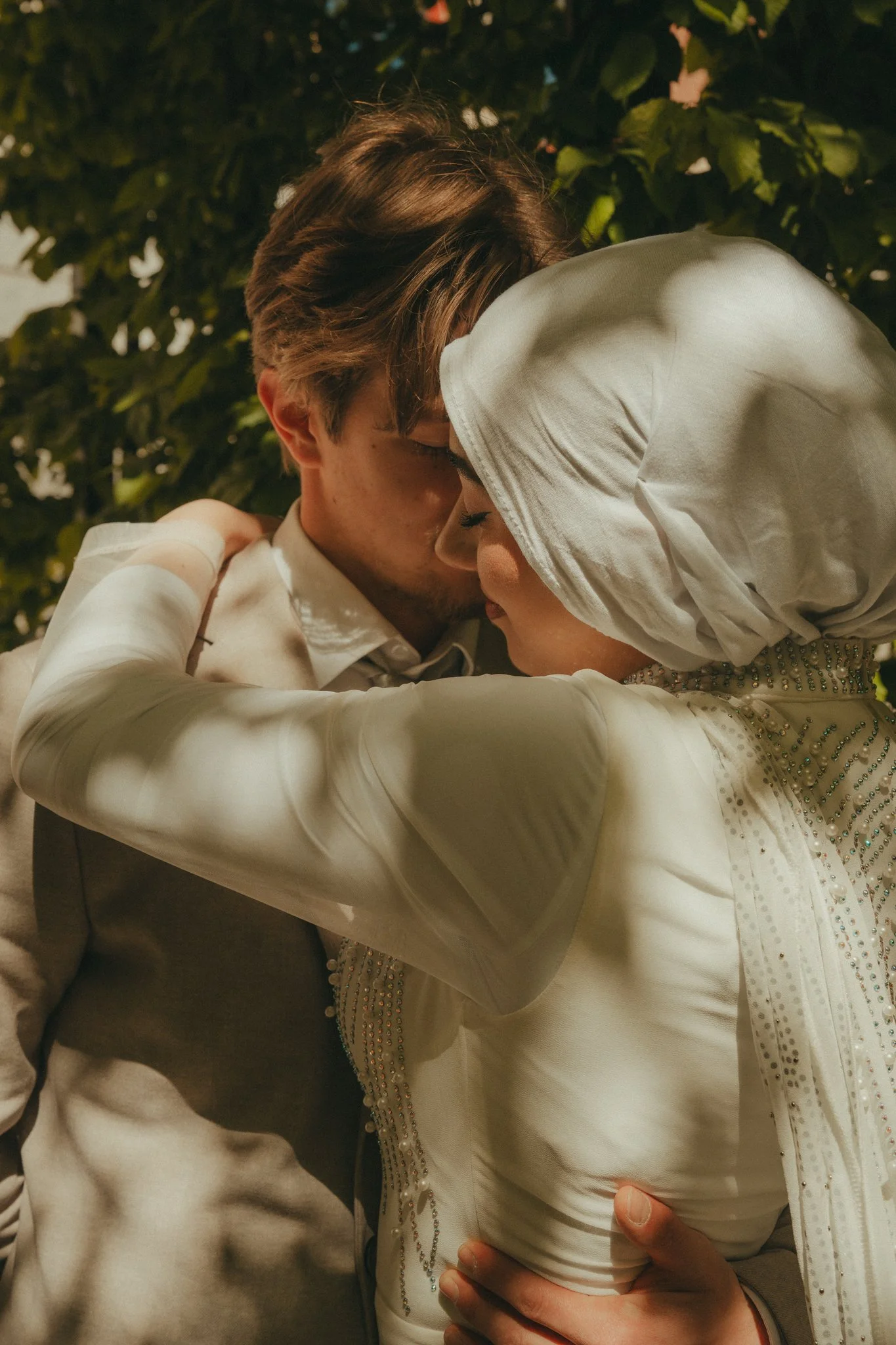 A couple sharing a kiss in an outdoor setting with green foliage in the background, dressed in formal attire.