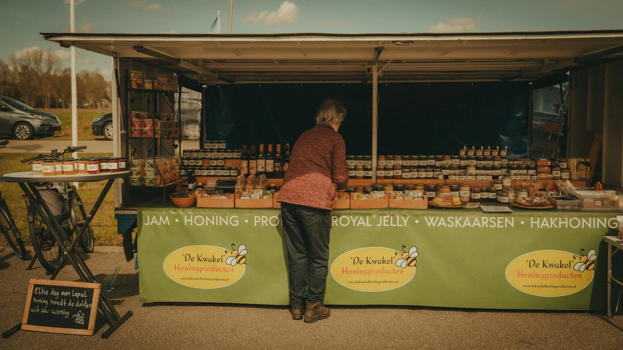 A woman at an outdoor honey stand with jars of honey, honeycomb, and bottles of honey syrup, under a canopy with signs for honey, jam, royal jelly, and Wackaeren products, in a field with parked cars in the background.
