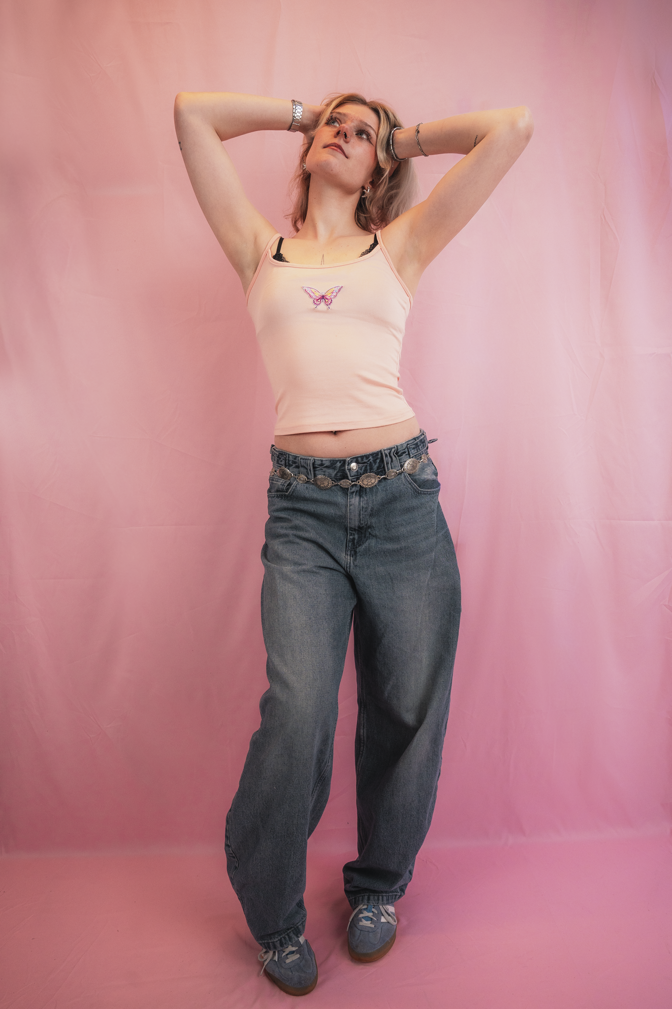 A young woman standing against a pink backdrop, wearing a peach tank top with a butterfly design, black bra, high-waisted jeans with a decorative belt, sneakers, and jewelry, with her hands behind her head looking upward.
