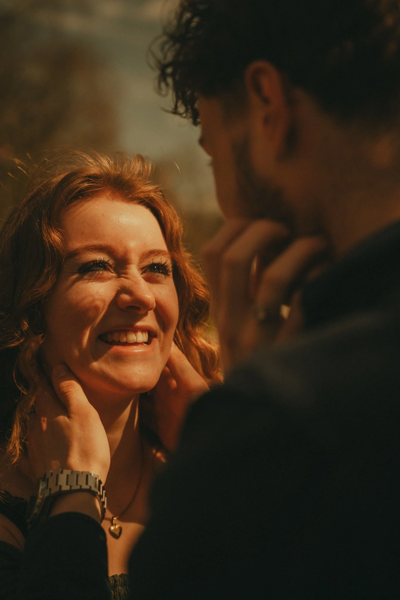 A woman with curly red hair smiling and looking up as a man leans in towards her, touching her face gently. The background is blurry with warm lighting.