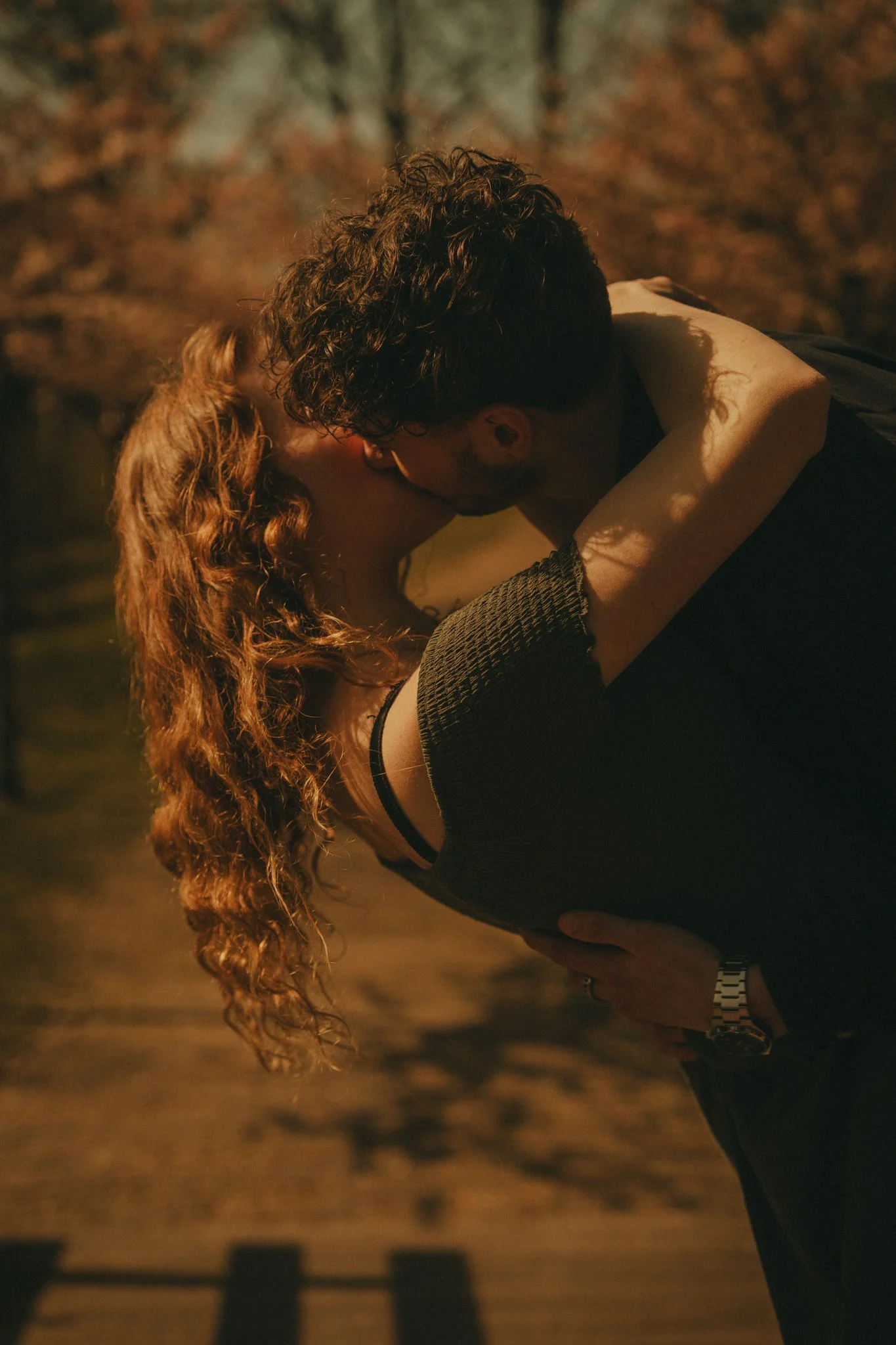 A couple kissing outdoors at night, with the woman having long, curly red hair and the man with dark, curly hair, both embracing each other.