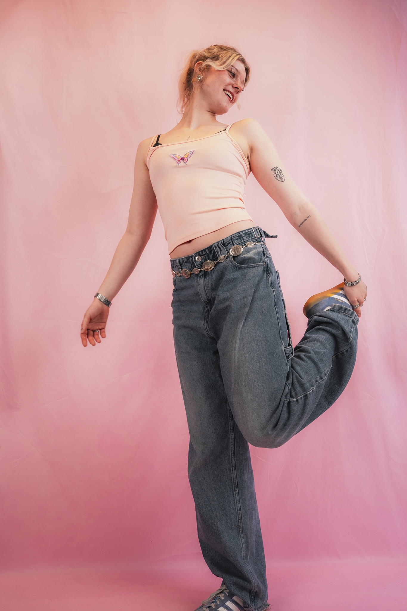 A young woman with blonde hair poses against a pink background. She is wearing a peach-colored tank top with a butterfly embroidery and dark jeans with a silver chain belt. She is smiling, looking to her right, with her left leg bent backwards and ho