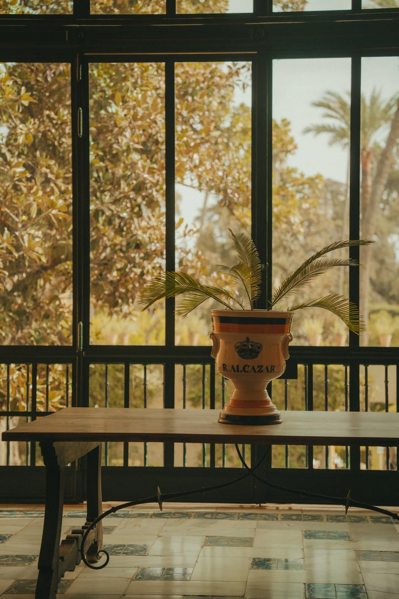 A decorative ceramic vase with a plant on a wooden table in front of a window, with a background of trees and palm trees outside.