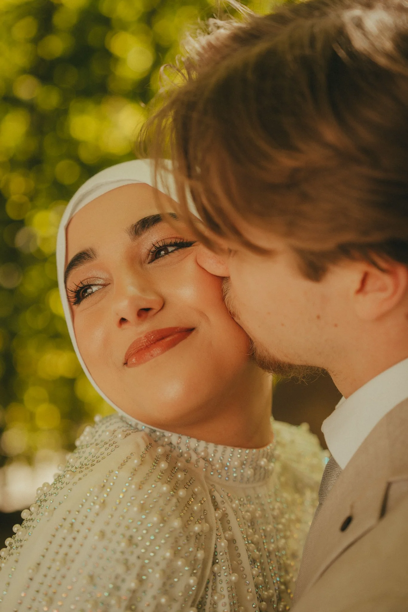 A woman wearing a white head covering and a dress with pearl and rhinestone embellishments smiling, as a man with brown hair leans in to kiss her on the cheek, with a background of blurred green foliage.