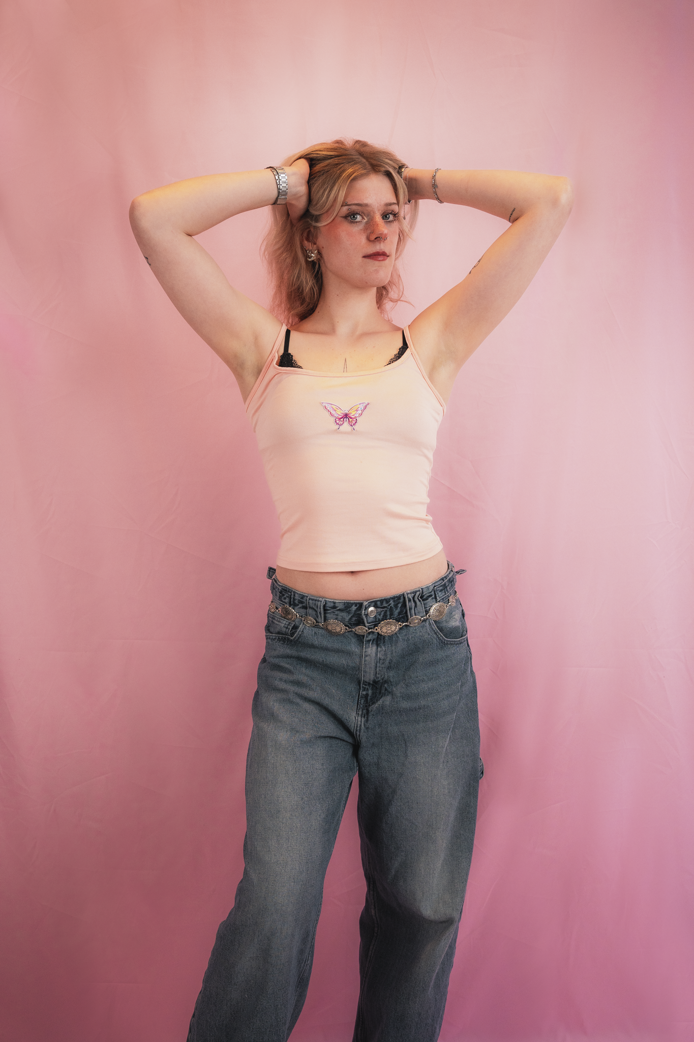 A woman with blonde wavy hair standing against a pink backdrop, wearing a pink tank top with a butterfly on it, black bra straps, and loose high-waisted jeans, with her hands behind her head.