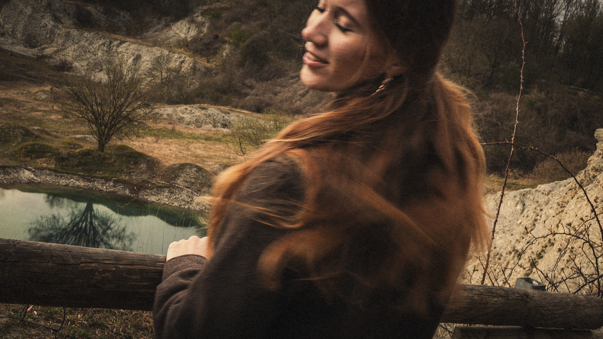 A woman with long reddish-brown hair smiling with her eyes closed, standing outdoors near a body of water surrounded by rocky terrain and sparse trees during what appears to be sunset.