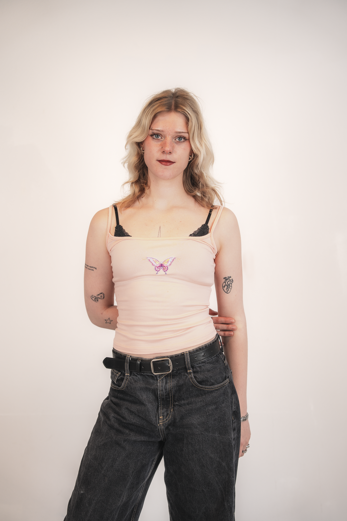 A young woman with wavy blonde hair and tattoos on her arms standing against a plain light-colored background. She is wearing a light pink tank top with a butterfly design, black lace straps, and dark denim jeans.