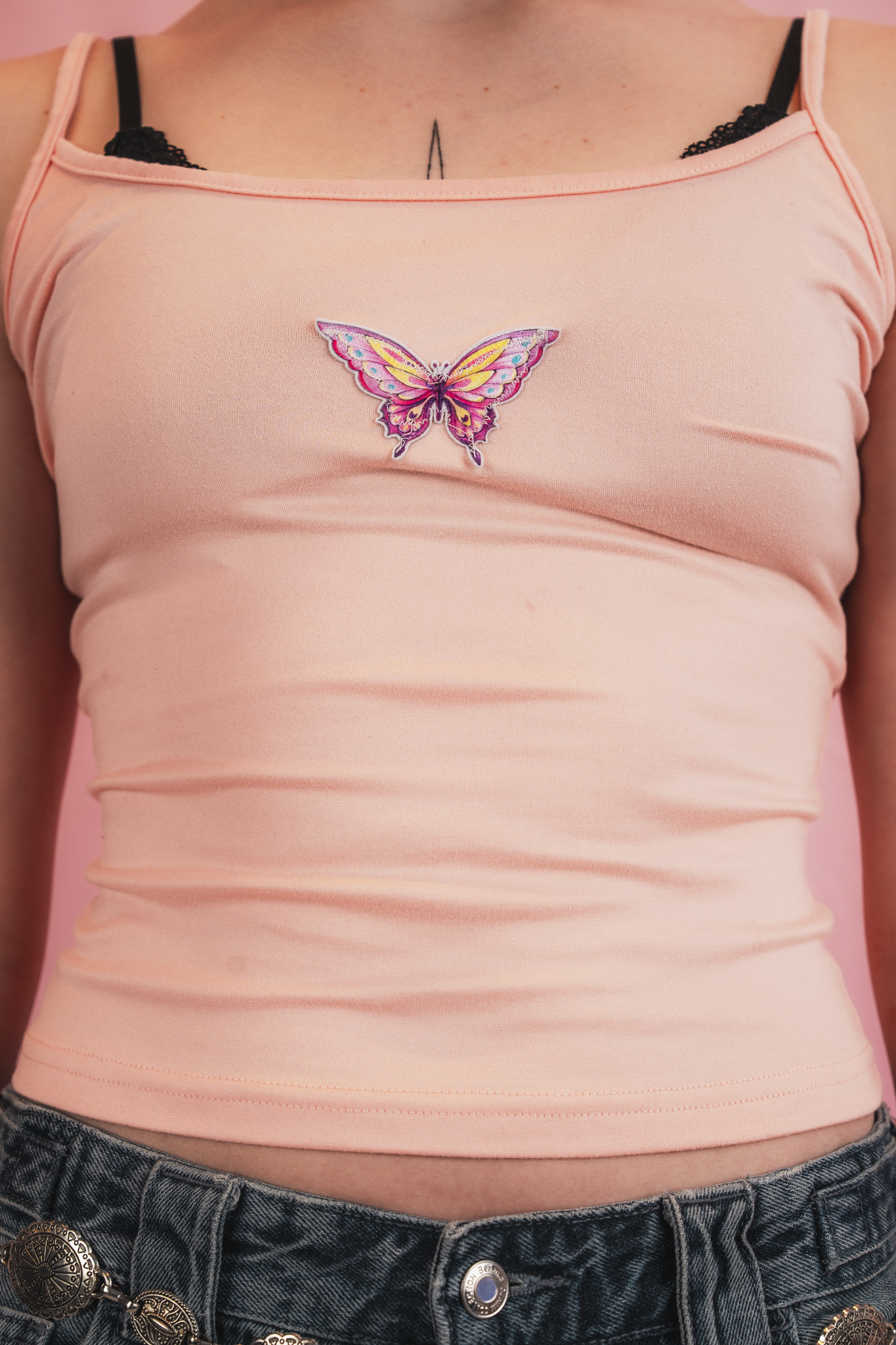 Close-up of a woman wearing a pink tank top with a butterfly patch in the center, black lace bra straps visible, and denim shorts with a decorative chain belt.