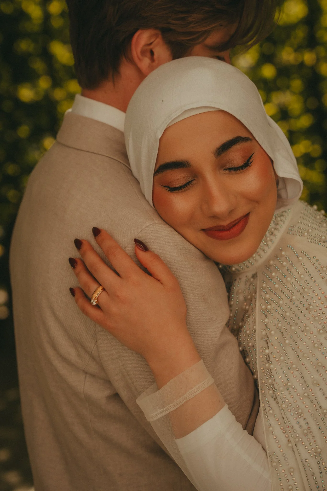 A woman with makeup and a white hijab hugging a man in beige suit and beige shirt, with a blurred green leaf background.