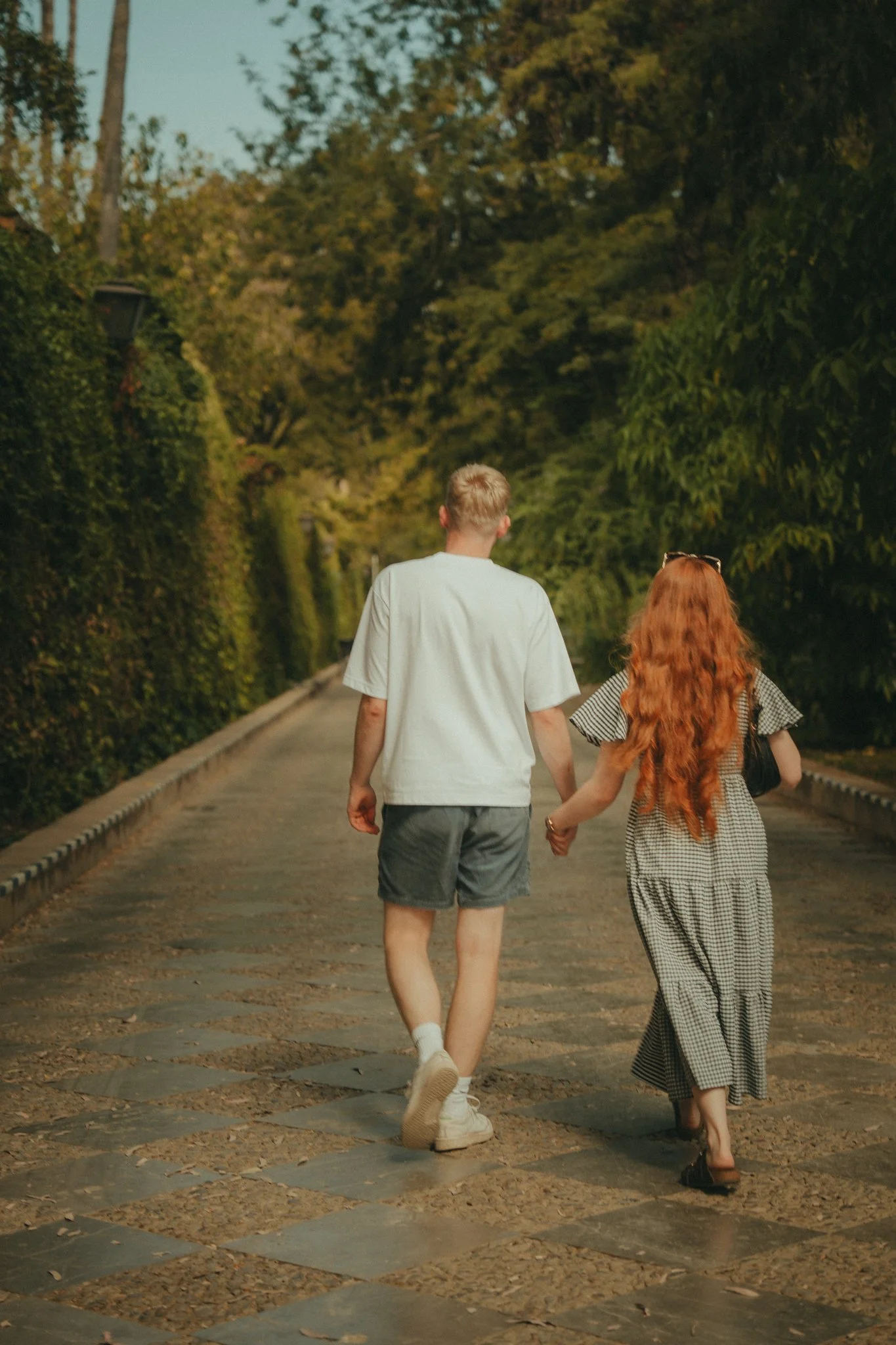 A young man and woman holding hands and walking away on a stone-paved path in a park surrounded by trees.