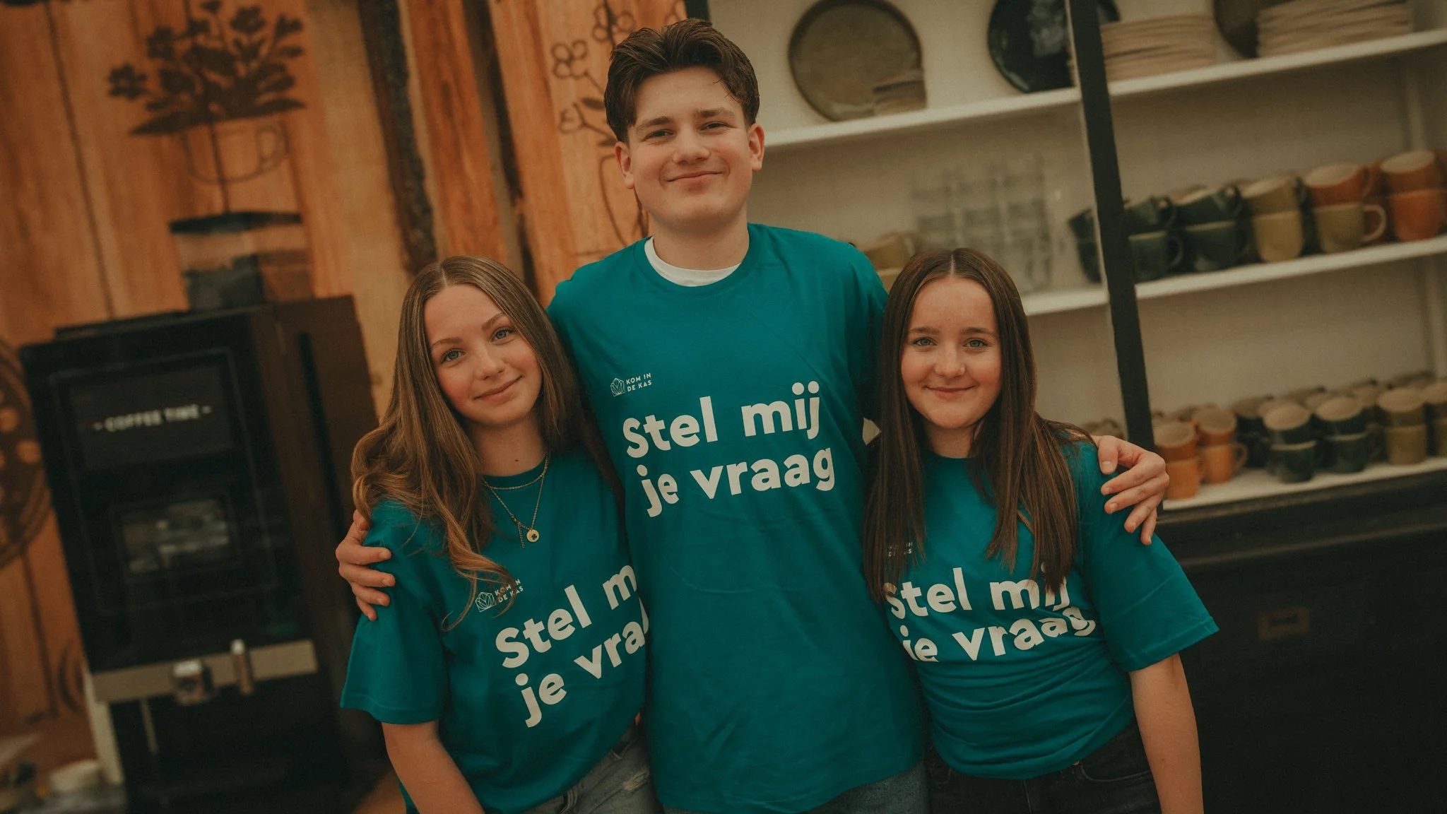 A group of three young people, two girls and one guy, smiling and standing close together indoors. They are wearing matching teal shirts with white text in Dutch that reads 'stel mij je vraag.' The background features a wooden wall and shelves with c