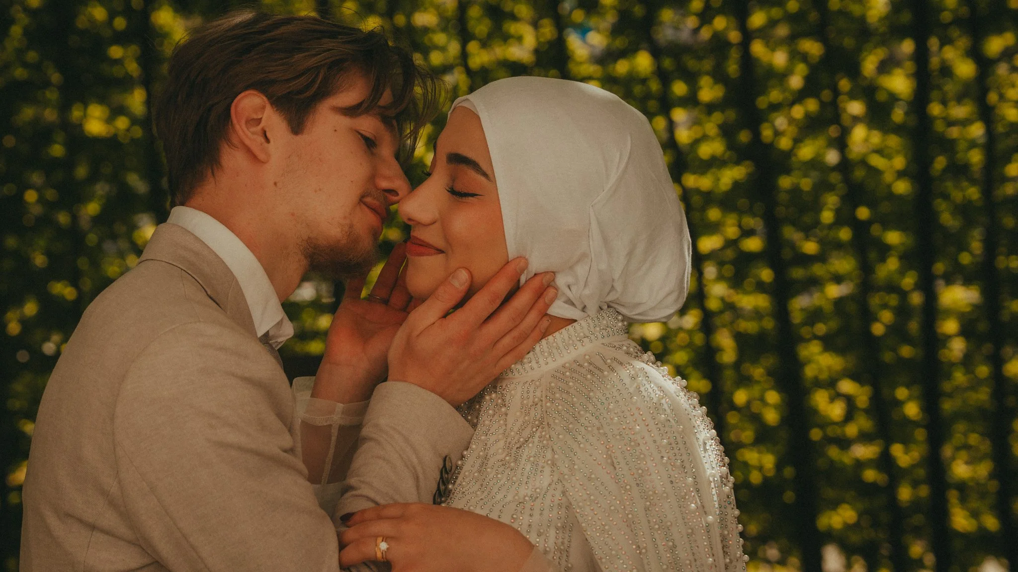 A young couple kissing outdoors amidst green trees, dressed in formal wedding attire.