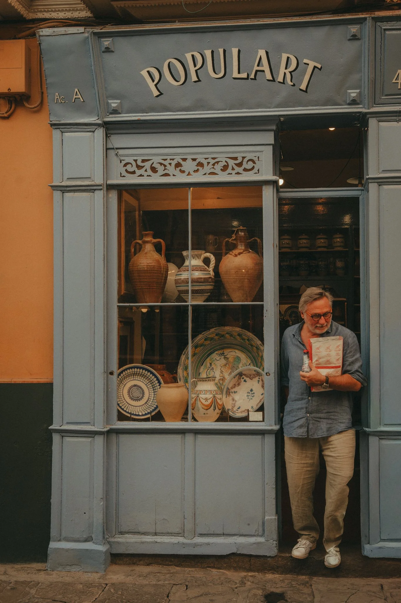 A man with glasses, gray hair, and a beard stands outside a storefront named "Popular". He is holding a menu or paper bag and leans against the doorframe. The storefront displays various pottery and decorative plates inside, with a large window showc