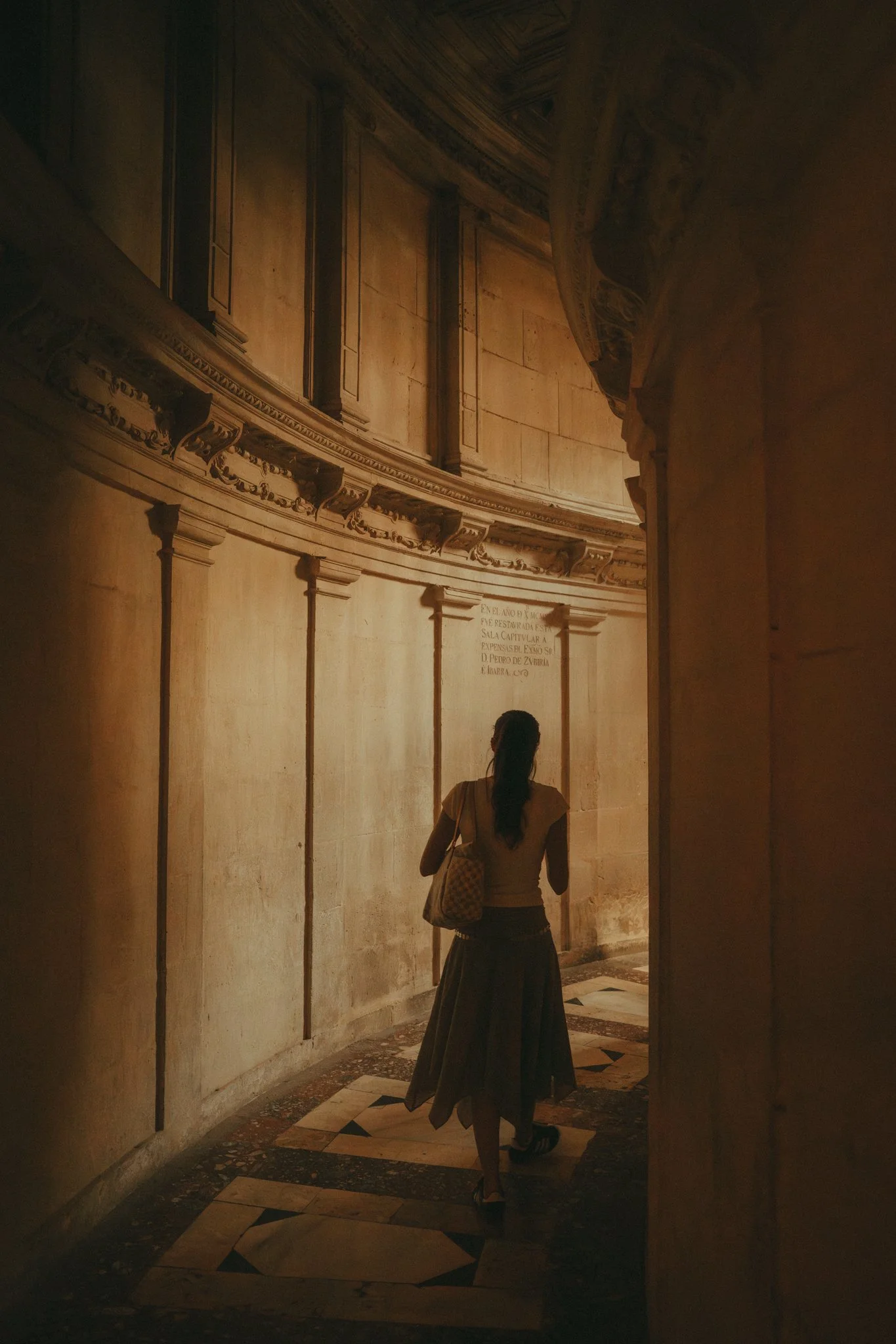 A woman with long dark hair wearing a black skirt, heels, and a beige top walking through a dimly lit historic corridor with ornate walls and a marble floor.