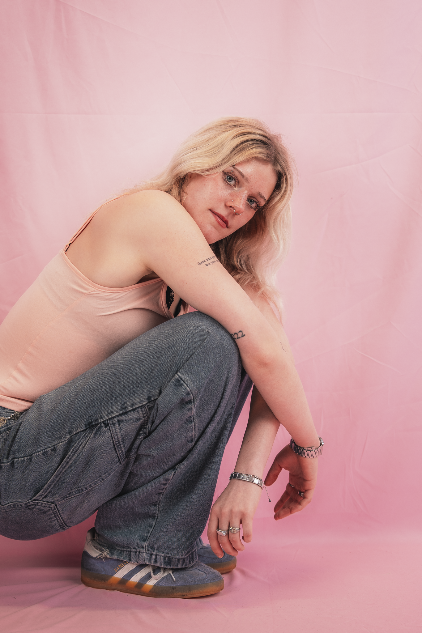 A person with long blonde hair, wearing a pink tank top, gray jeans, gray sneakers, and jewelry, sitting on the floor against a pink backdrop.
