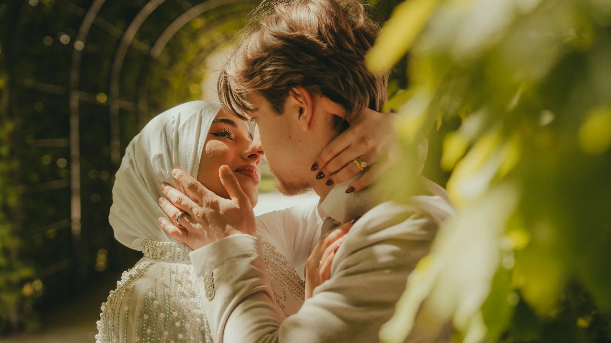 A couple face each other closely, holding each other's faces, in a romantic moment outdoors, surrounded by greenery.