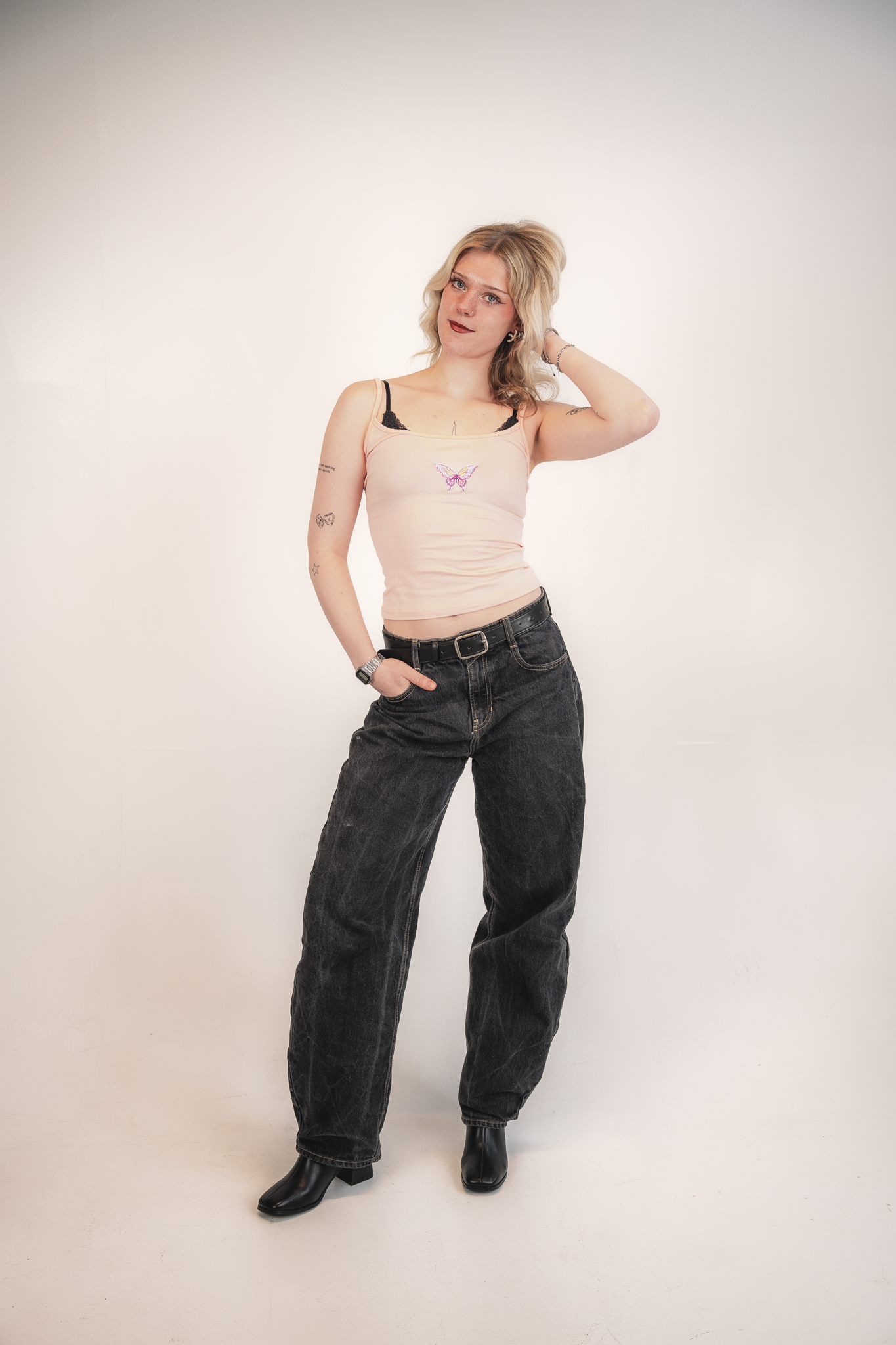 A woman with blonde hair styled in loose waves, wearing a beige tank top with a butterfly graphic on the chest, black jeans, and black ankle boots, standing against a plain white background.