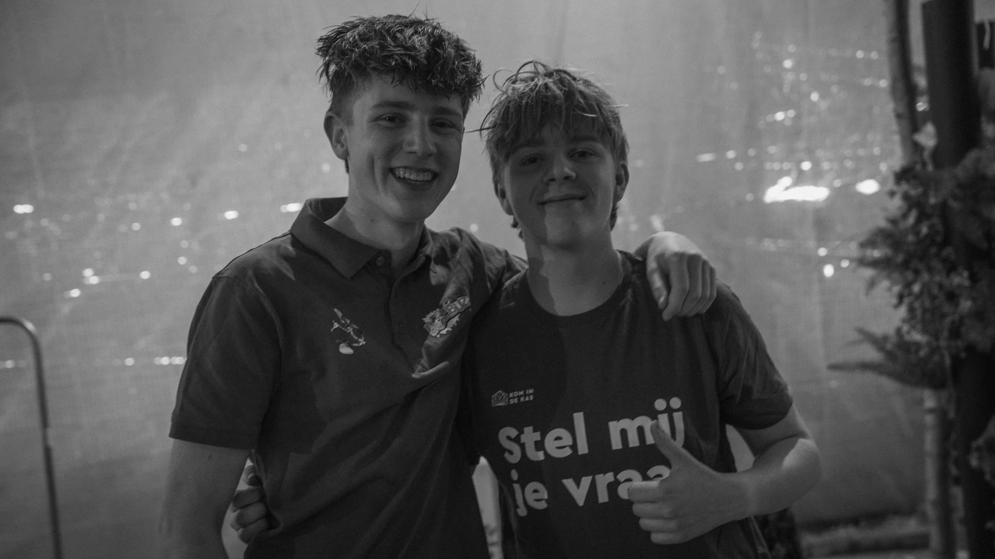 Two teenage boys with messy hair smiling and posing together, one with arm around the other, in front of a textured background.