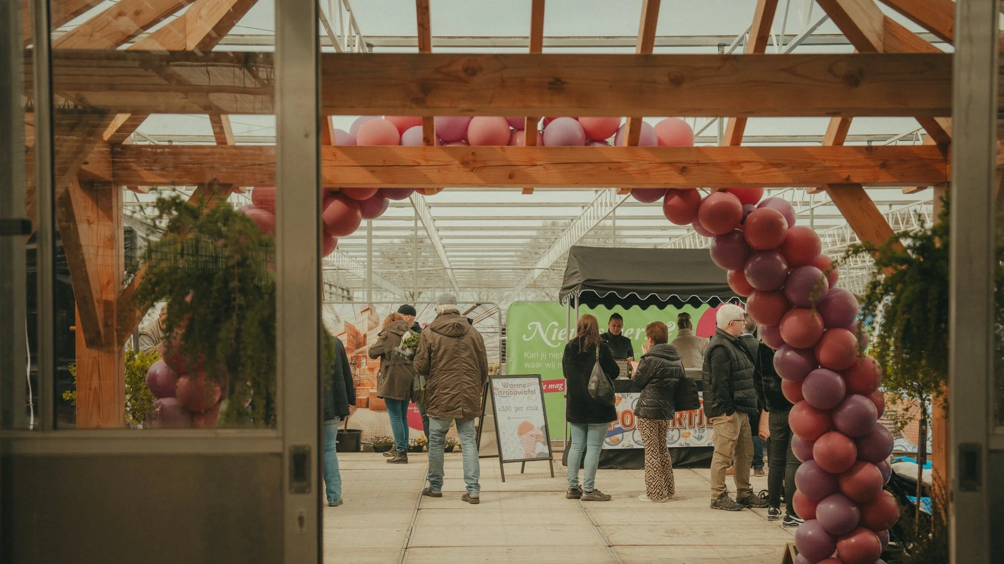 View through a window showing people shopping at a farmers' market with a decorative balloon arch in the foreground.