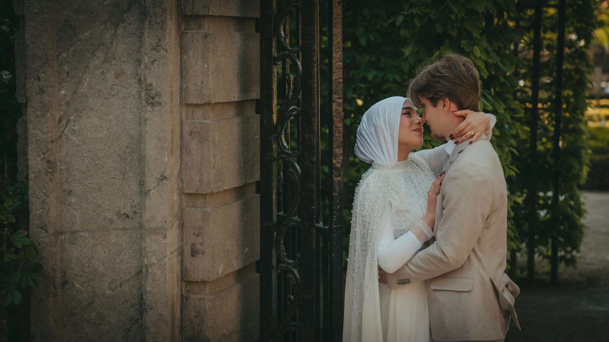 A couple embracing outside near a stone wall with a decorative iron gate, surrounded by trees and greenery.