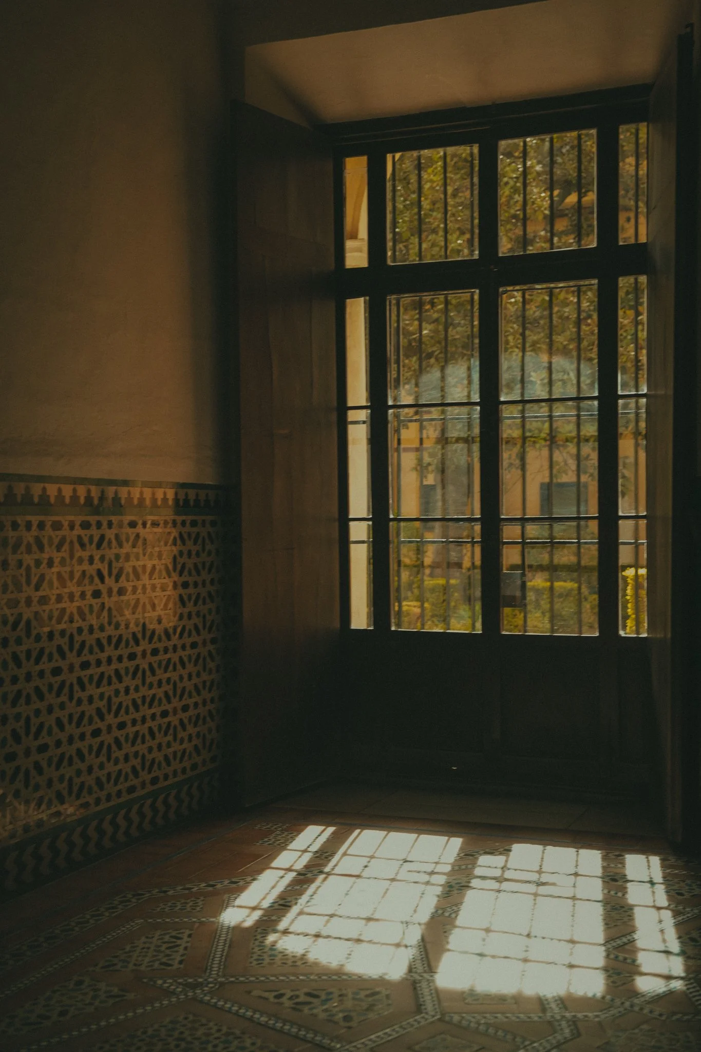 Sunlight coming through a large barred window, casting geometric shadows on the tiled floor inside a room.