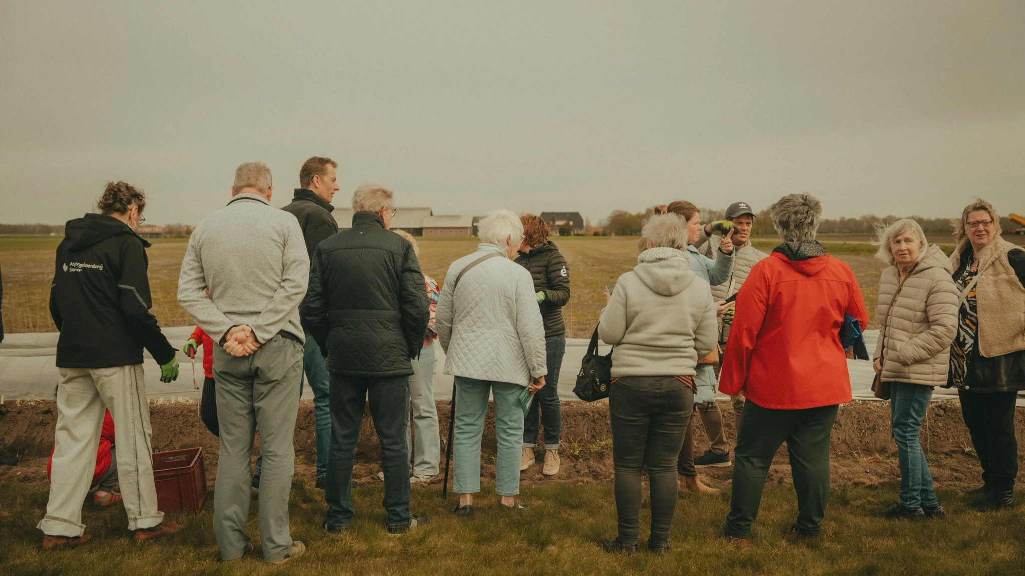 Group of people outdoors on a cloudy day, gathered around looking at something on the ground with farm buildings in the background.