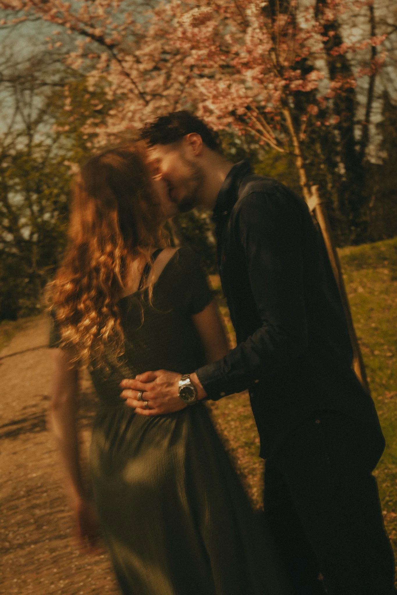 A couple standing close together outdoors during evening, about to kiss, with cherry blossom trees in full bloom in the background.