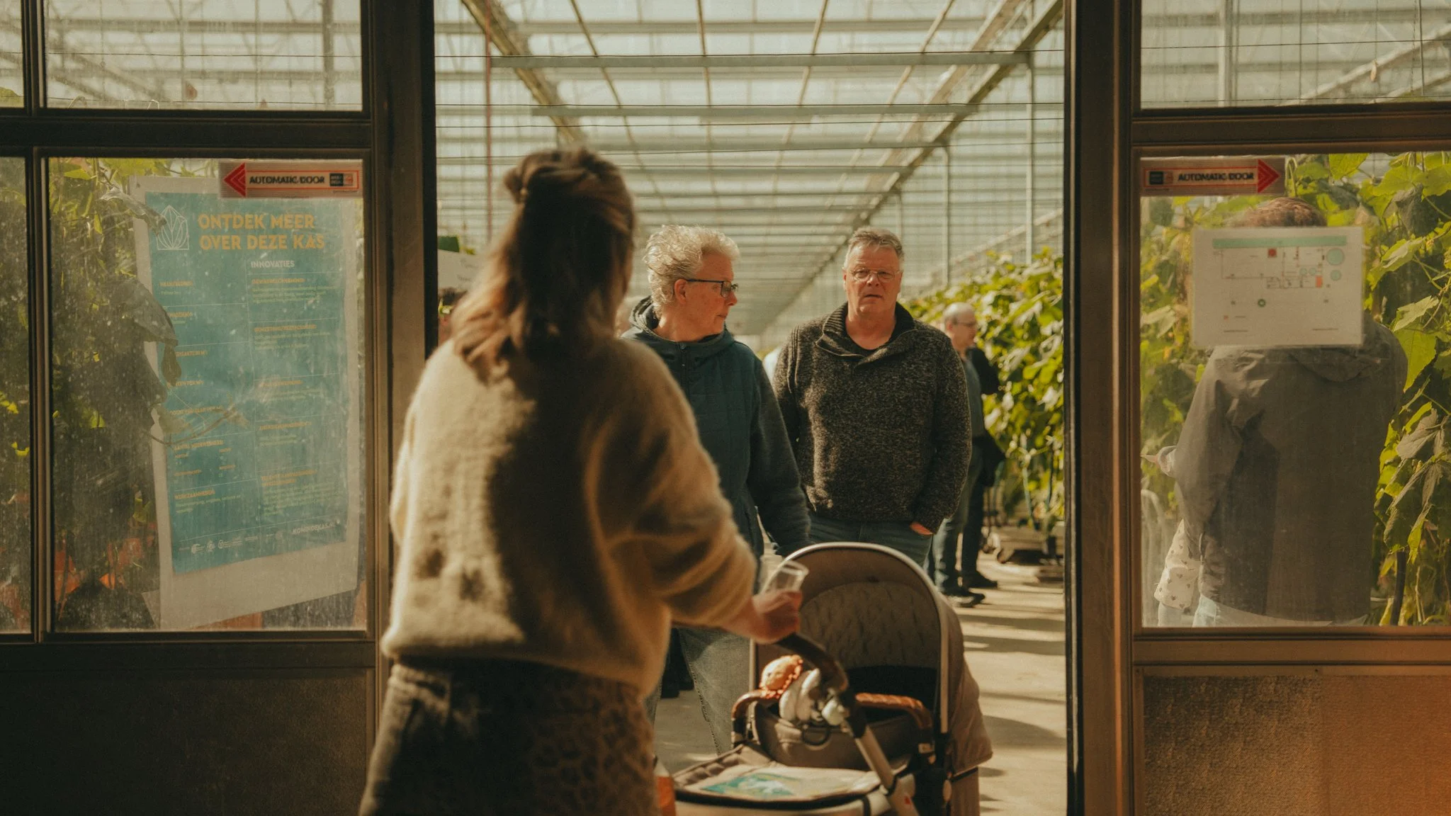 View of people inside a glass greenhouse or plant nursery, with sunlight coming through the glass ceiling, and others walking or standing outside.