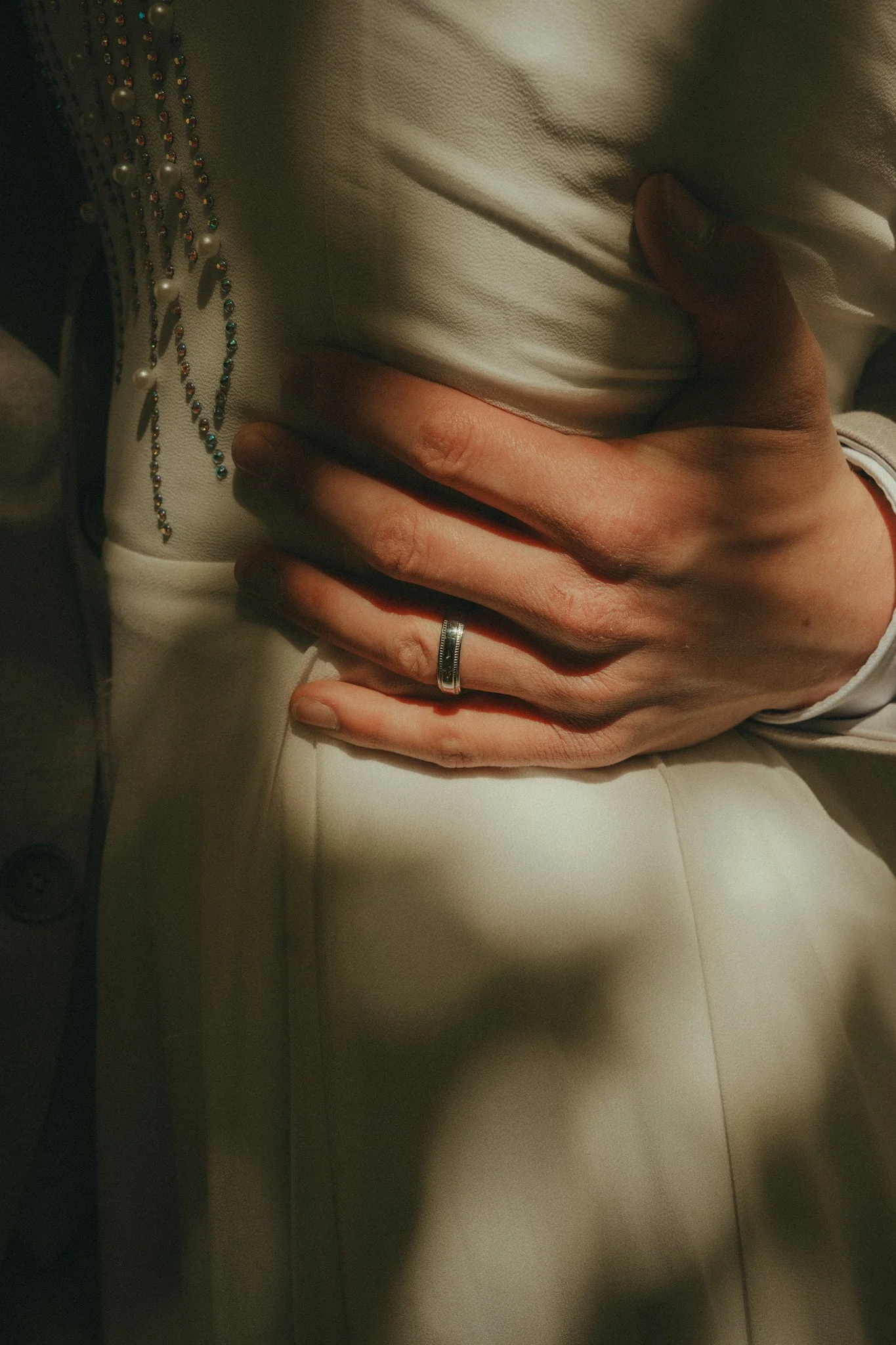 Close-up of a person's hand resting on their thigh, wearing a silver ring, with a beige or cream-colored outfit and a beaded necklace with pearls and colorful beads.