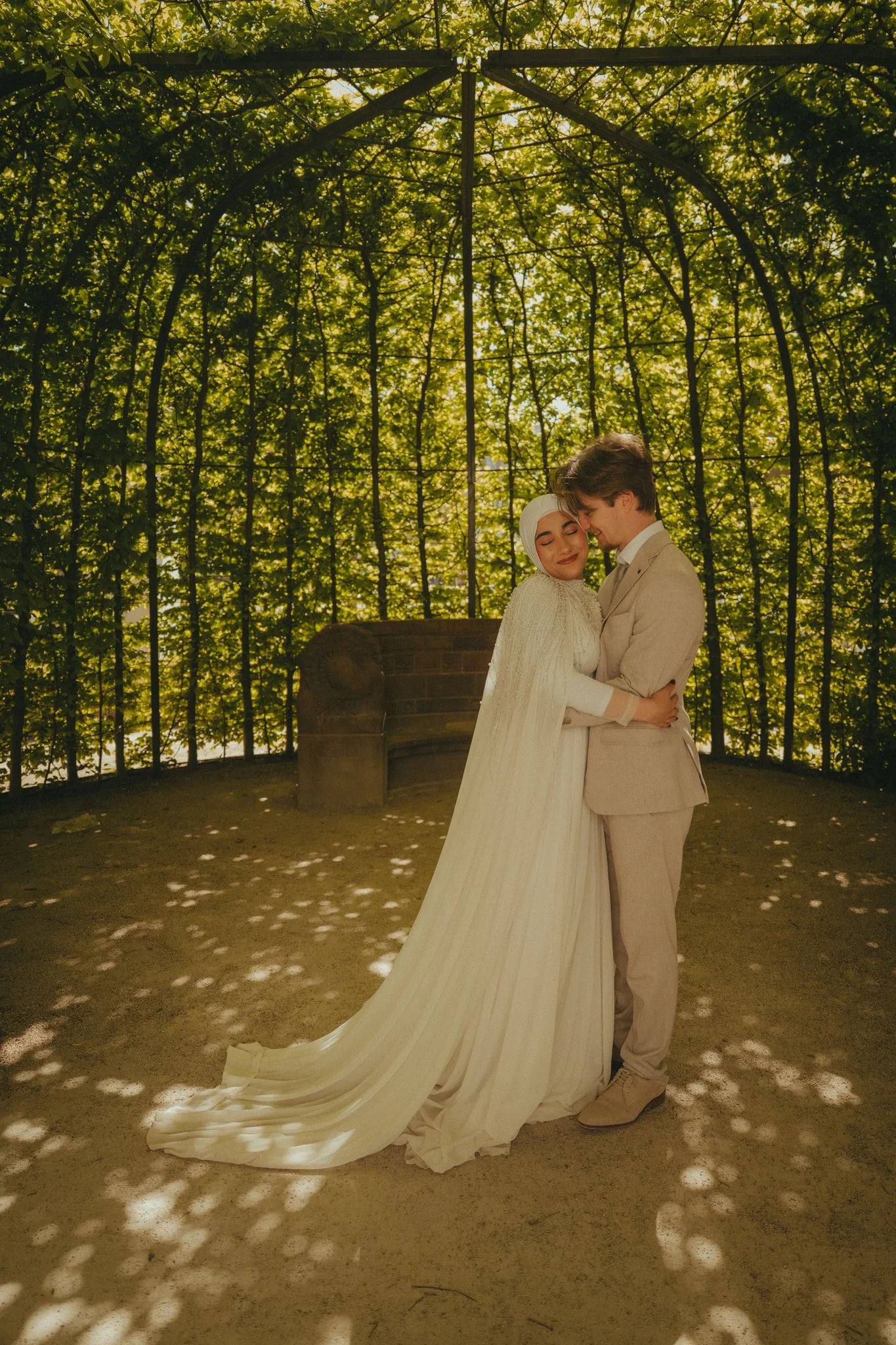 A bride and groom embrace in front of a decorative wrought iron and leafy background, sunlight filtering through the trees, with a brick structure behind them.