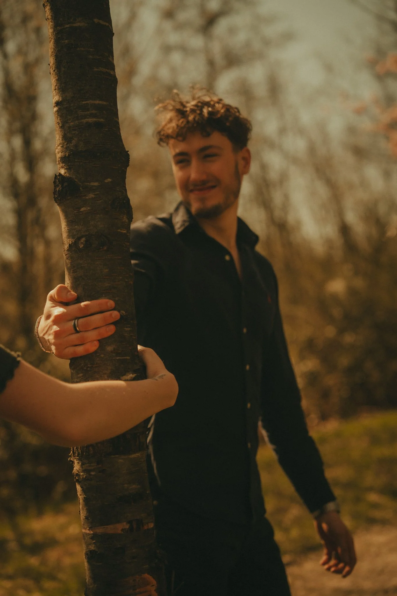 A young man with curly hair and a beard, dressed in black, standing outdoors behind a tree, smiling and looking to the side, in a forest setting with warm lighting.