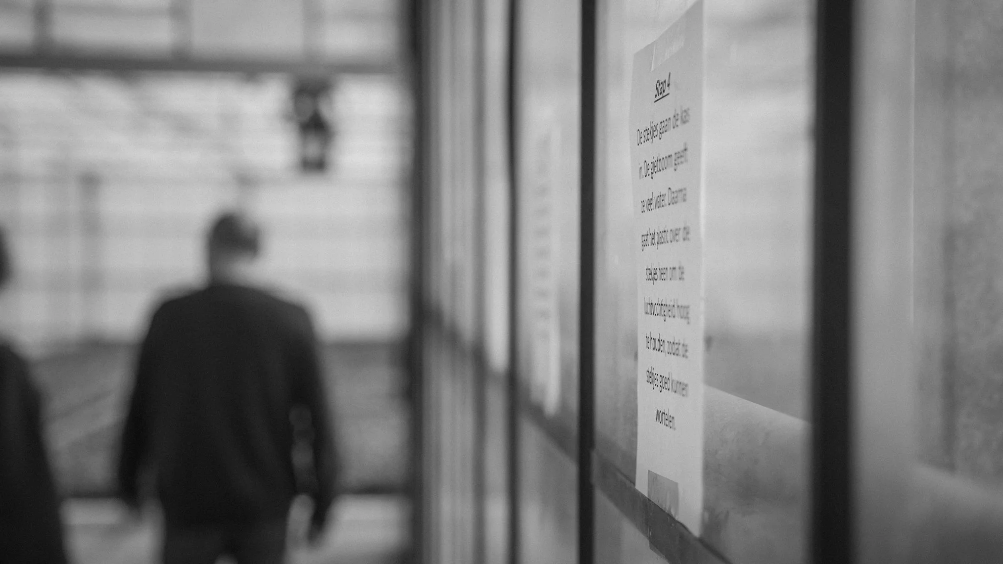 A blurry, black-and-white photo of a man walking in a corridor. A wall with informational posters or signs is visible on the right side of the image.