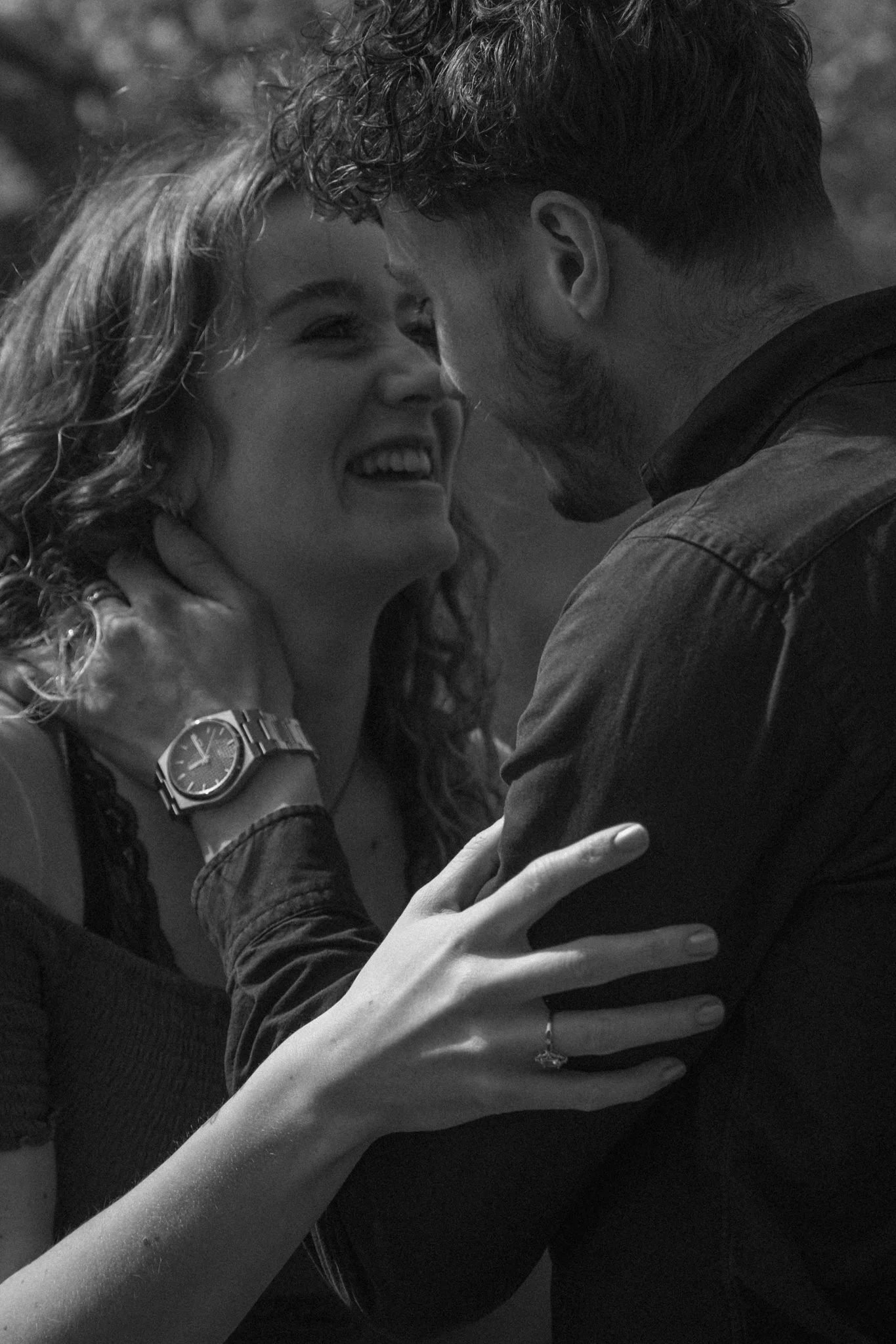 A black and white photo of a couple touching foreheads and smiling. The woman has curly hair, a watch on her left wrist, and a ring on her left ring finger. The man has short hair and a beard, and is wearing a dark jacket.