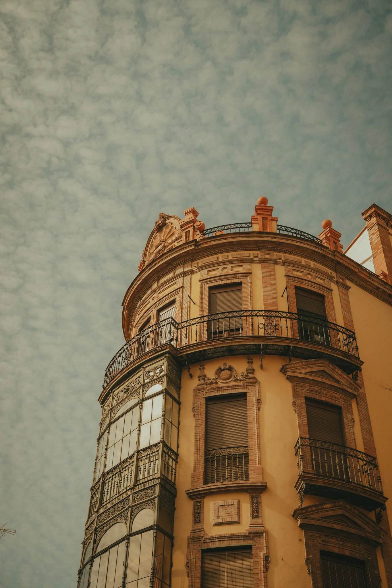 A tall, ornate building with multiple floors and curved balconies, topped with decorative elements and columns, under a partly cloudy sky.