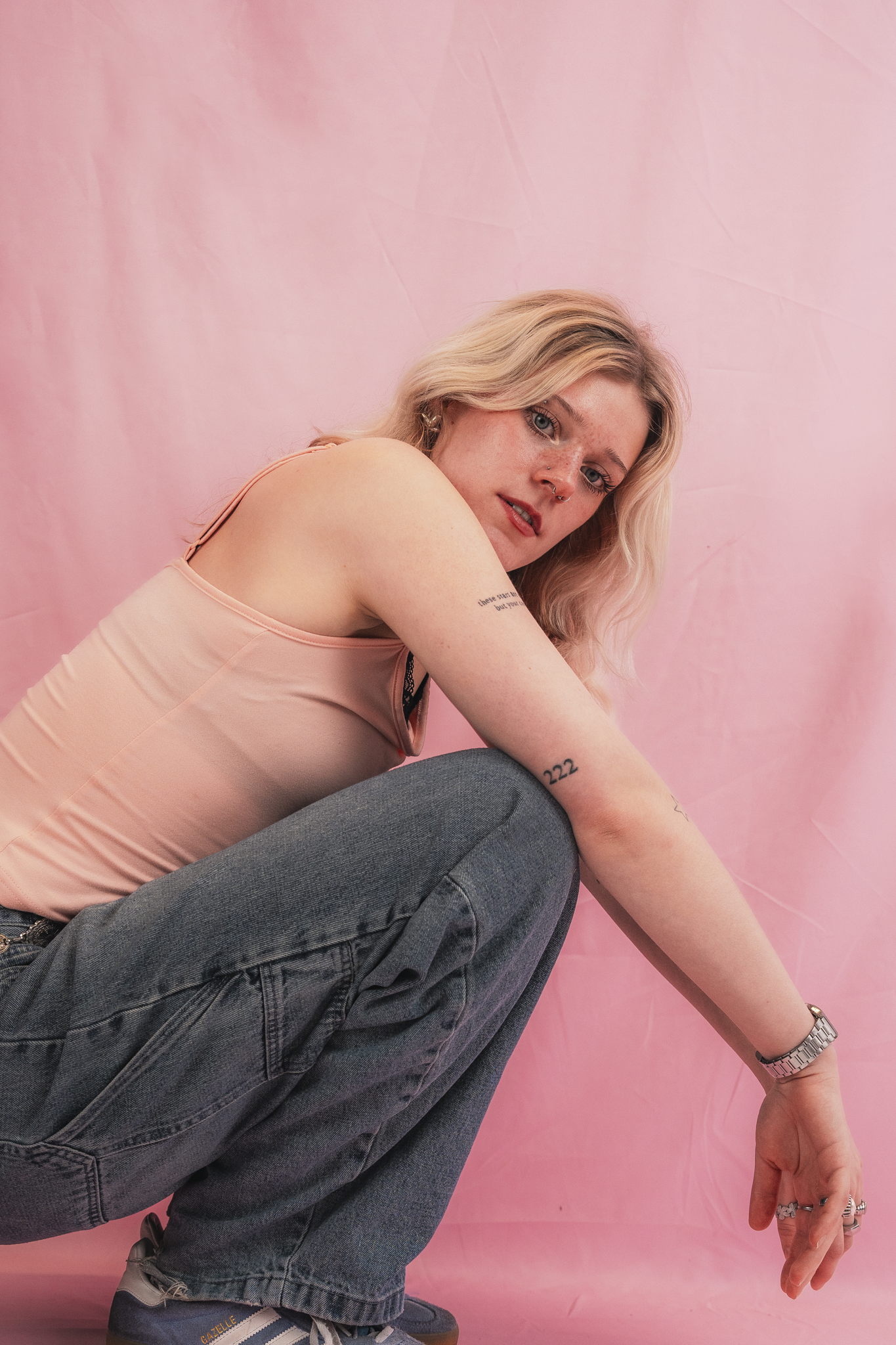 Young woman with blonde wavy hair, light skin, and tattoos, wearing a pink tank top, blue jeans, and sneakers, sitting against a pink background.