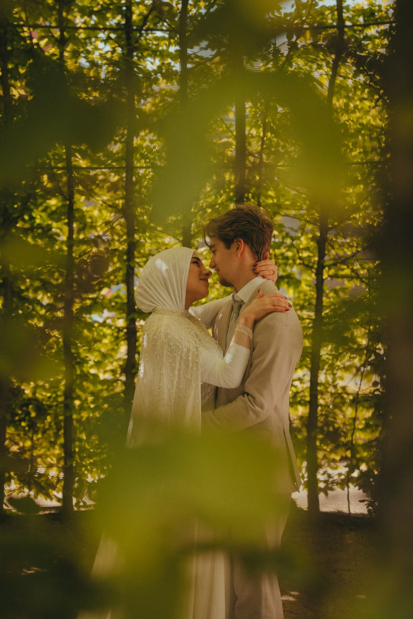 A couple embracing each other and smiling, standing close in a lush, green outdoor setting with sunlight filtering through trees.