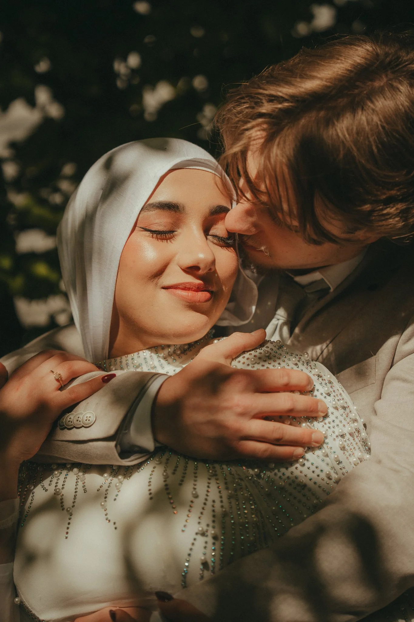A couple embracing at night, with the woman wearing a rhinestone embellished white dress and a headscarf, smiling with closed eyes, and the man dressed in a suit leaning close to her.