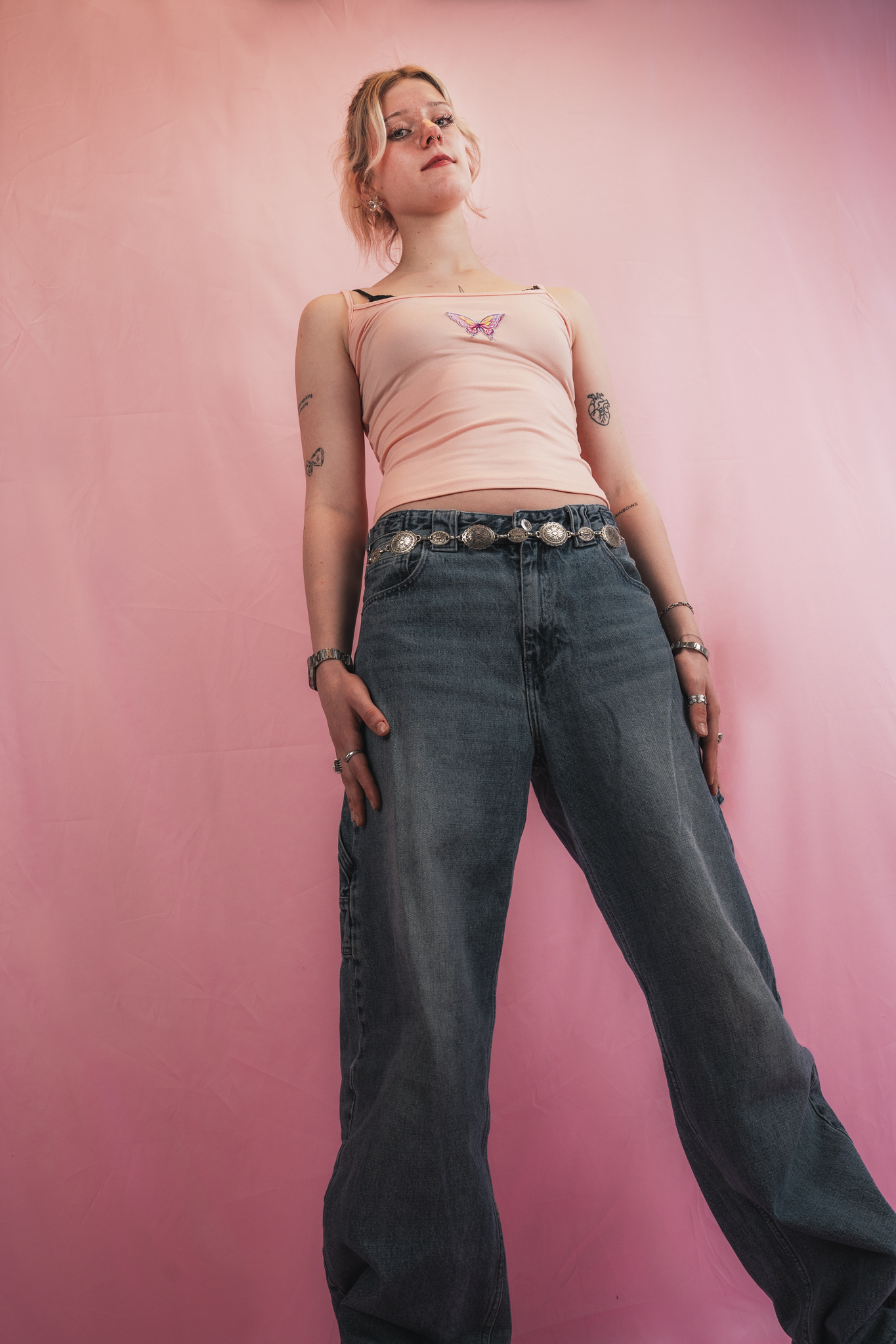 A young woman with short blonde hair, freckles, and tattoos on her arms, wearing a light pink tank top with a butterfly patch and high-waisted loose-fitting jeans with a large silver belt, standing against a pink backdrop.