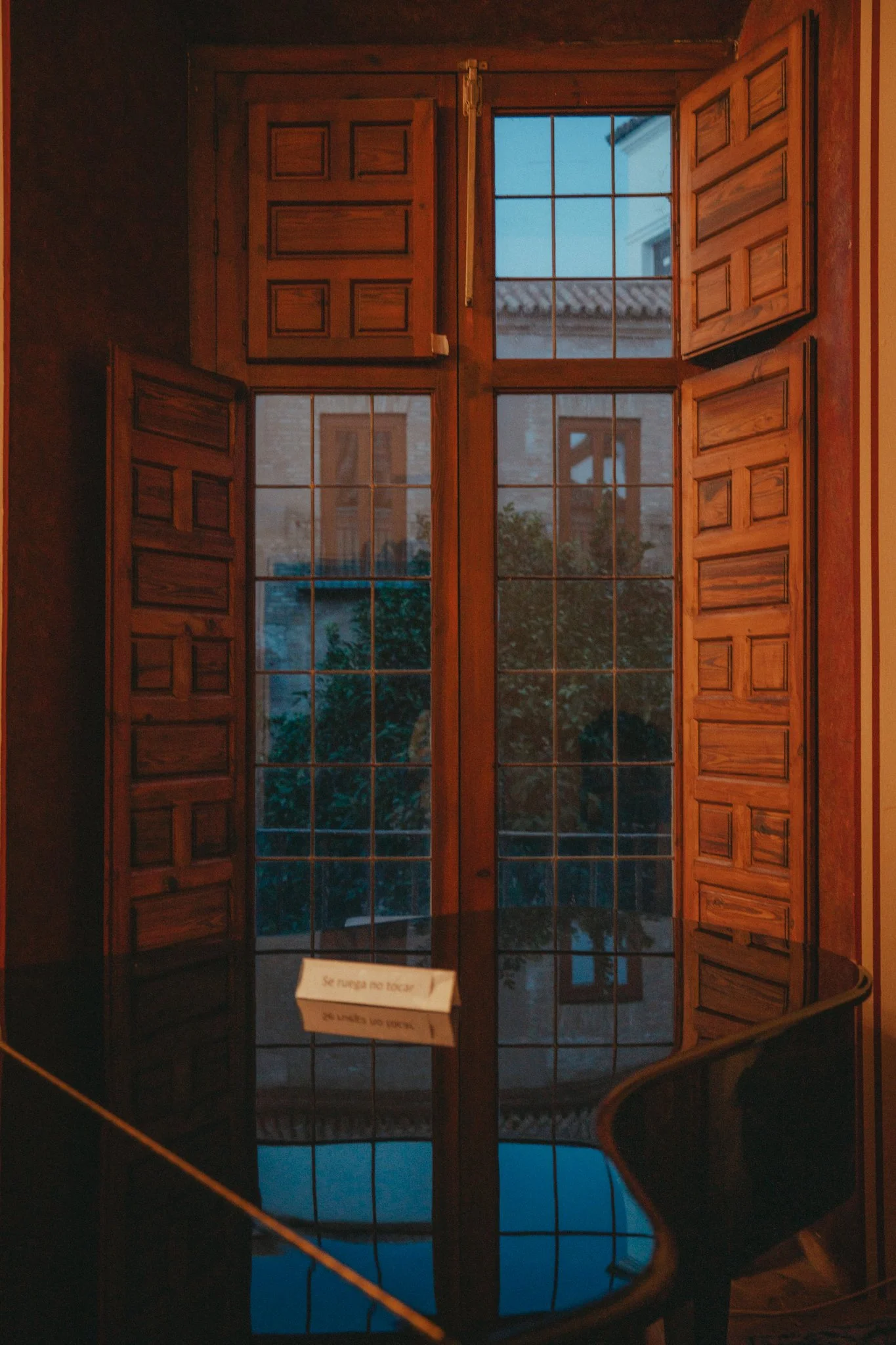 A wooden window with shutters, partly opened, revealing a view outside of a building with a tiled roof and a tree in the distance, with reflections visible on a shiny surface below.
