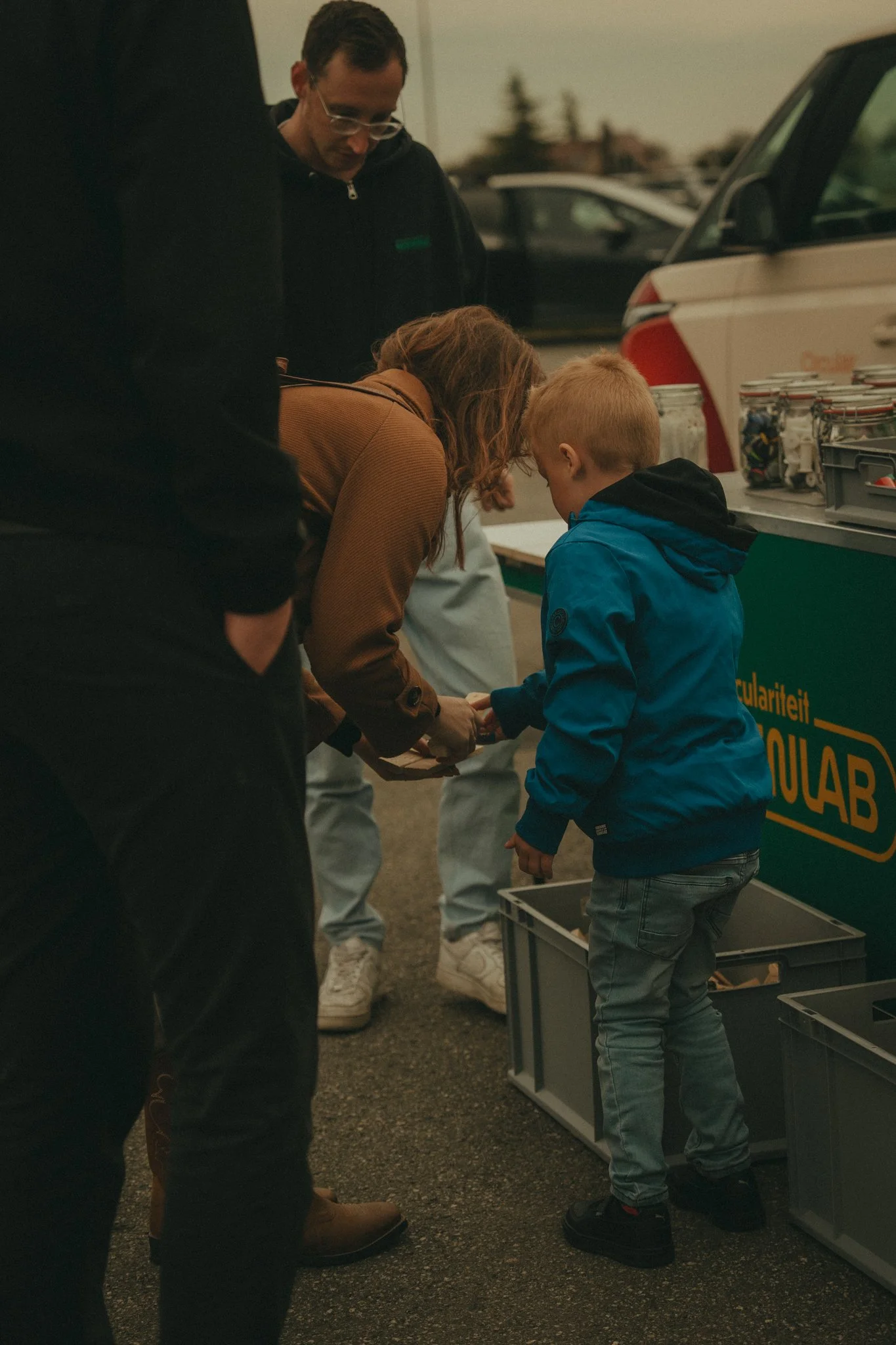 People gather around a display table at an outdoor event, possibly a recycling or community initiative, with several open containers and a vehicle with a logo in the background.