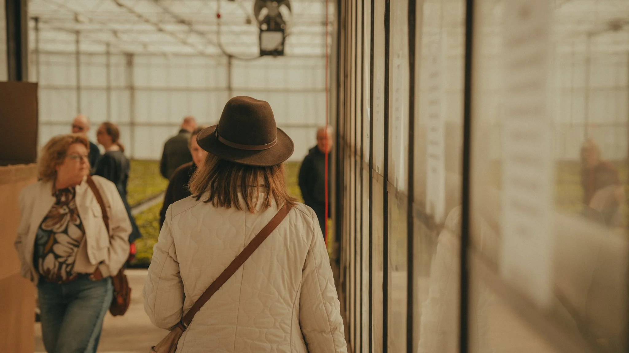 A woman with shoulder-length hair, wearing a brown hat and white jacket, stands in front of a window display with multiple documents posted on the wall. Several people are walking and conversing nearby inside a building with a high ceiling.
