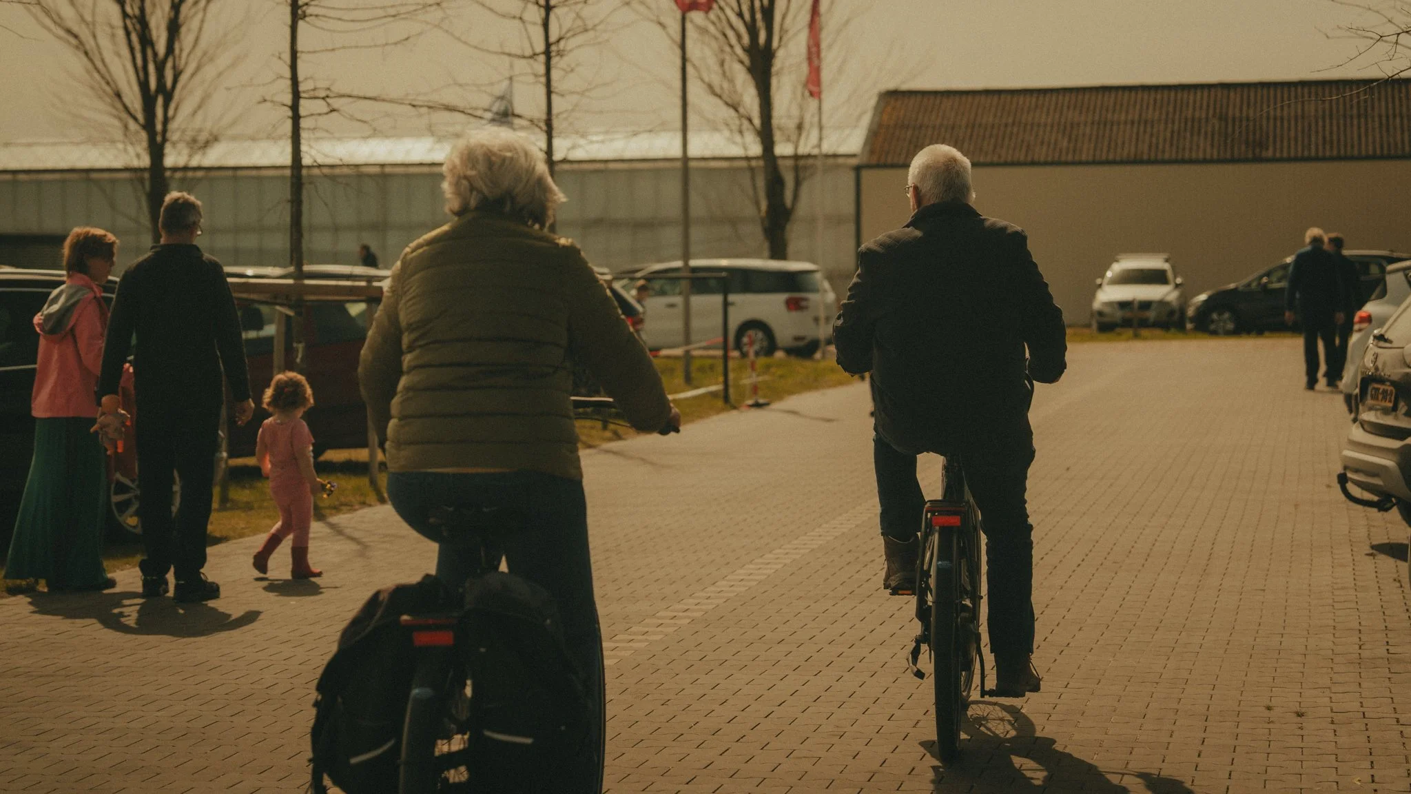 People in a parking lot, some riding bikes, and others standing or walking, during late afternoon or early evening.