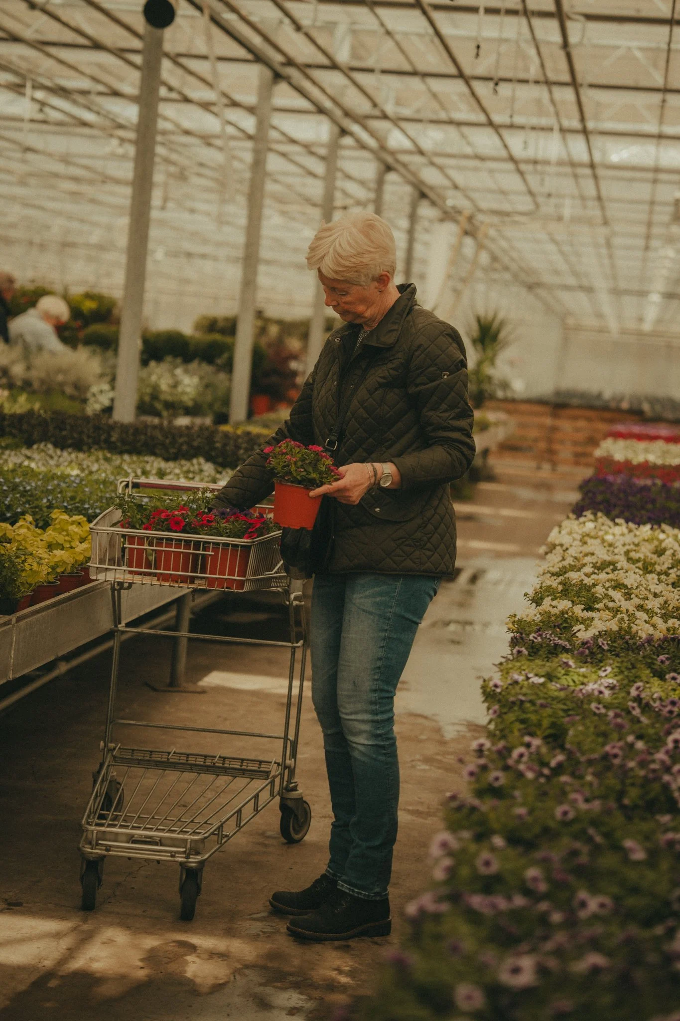 A woman with short blonde hair in a quilted black jacket and jeans shopping for flowers in a greenhouse, holding a potted plant and pushing a shopping cart. The background shows rows of plants and flowers.
