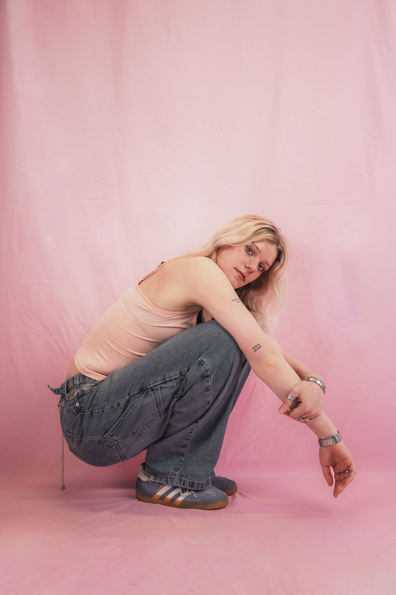 A person with blonde hair and tattoos, wearing a peach tank top and oversized jeans, balancing on one foot on a pink background.