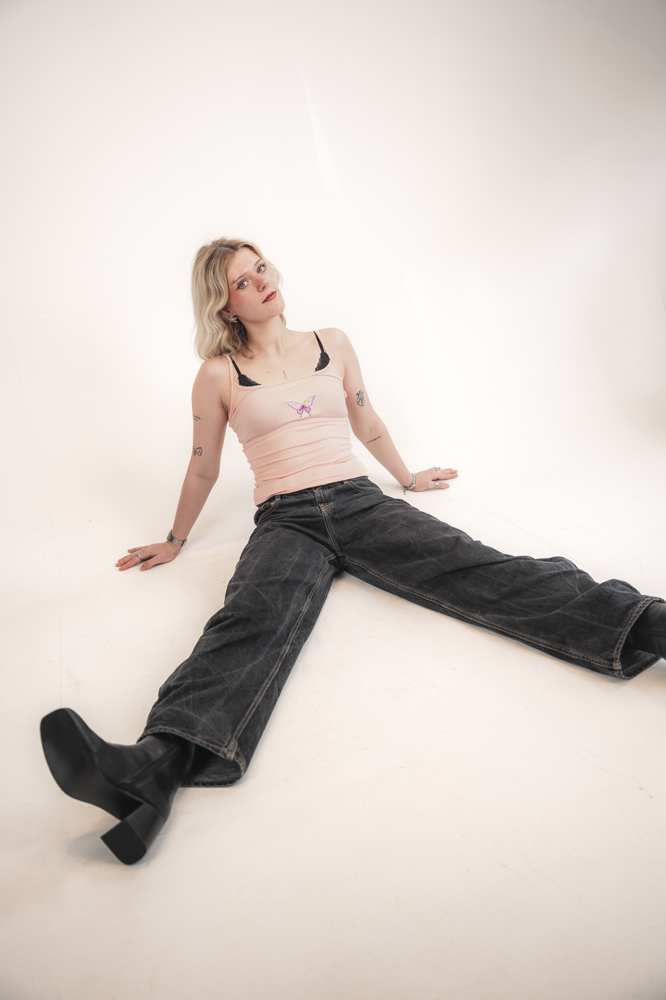 A young woman sitting on the floor against a plain white background, wearing a pink tank top with black lace details, black jeans, and black boots. She has blonde wavy hair, visible tattoos on her arms, and is looking slightly off-camera with a neutr