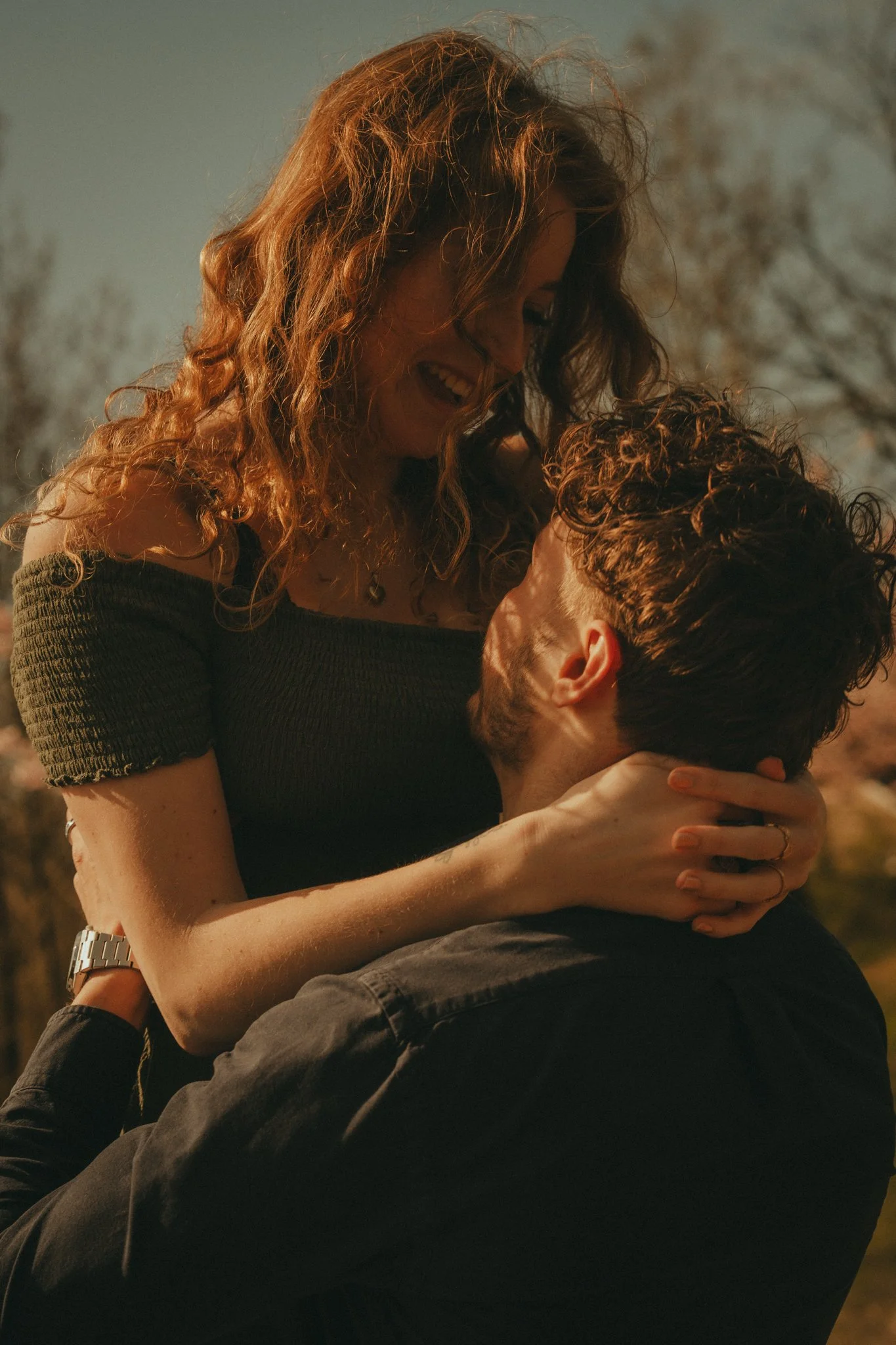 A woman with curly red hair smiling and leaning on a man's shoulder, outdoors with trees in the background.