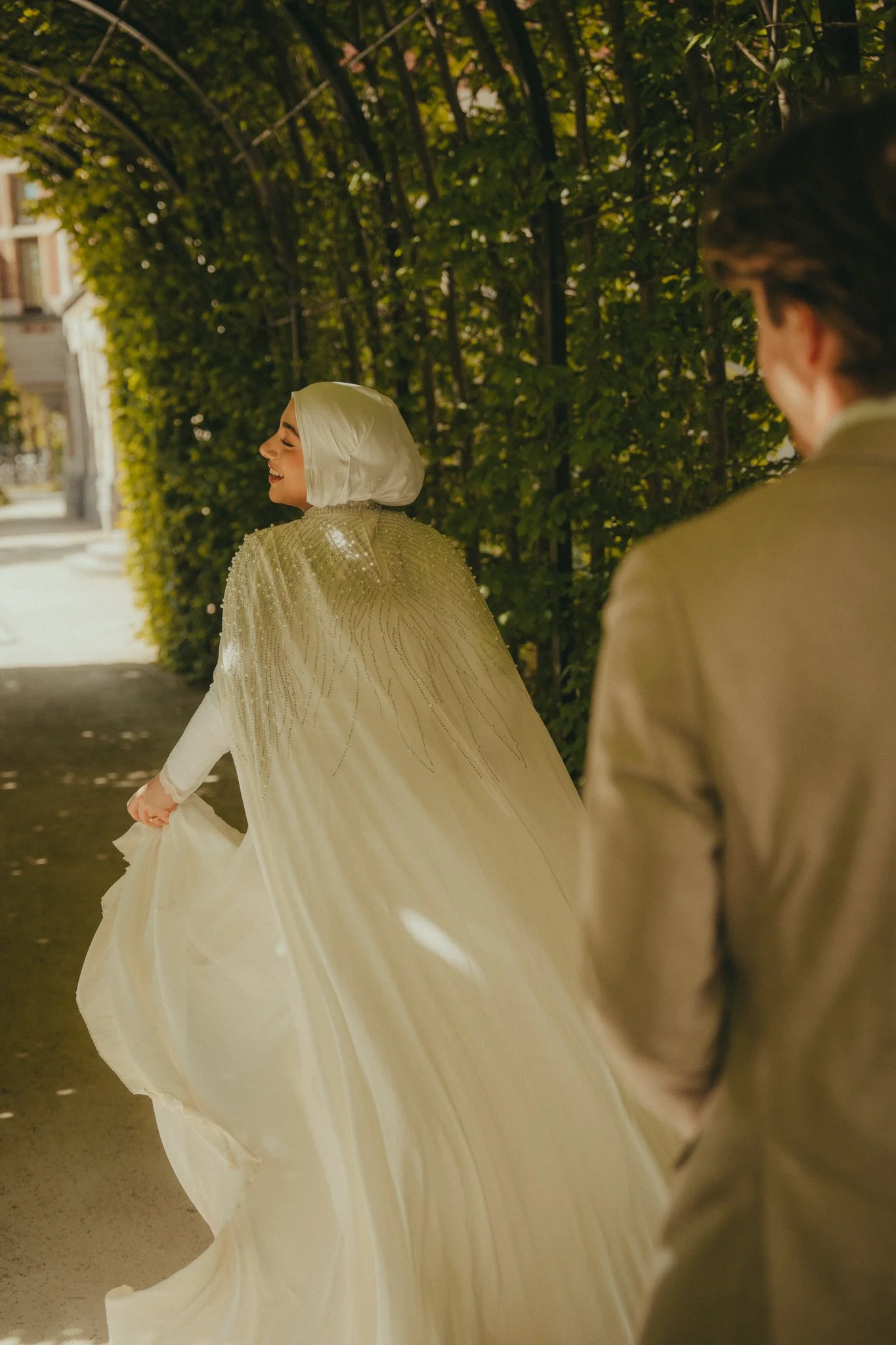 A woman wearing a white hijab and a cream-colored, embellished gown is smiling and turning her head to the left while holding her dress. A man in a beige jacket is seen from behind on the right side of the image. They are outdoors, next to a green he